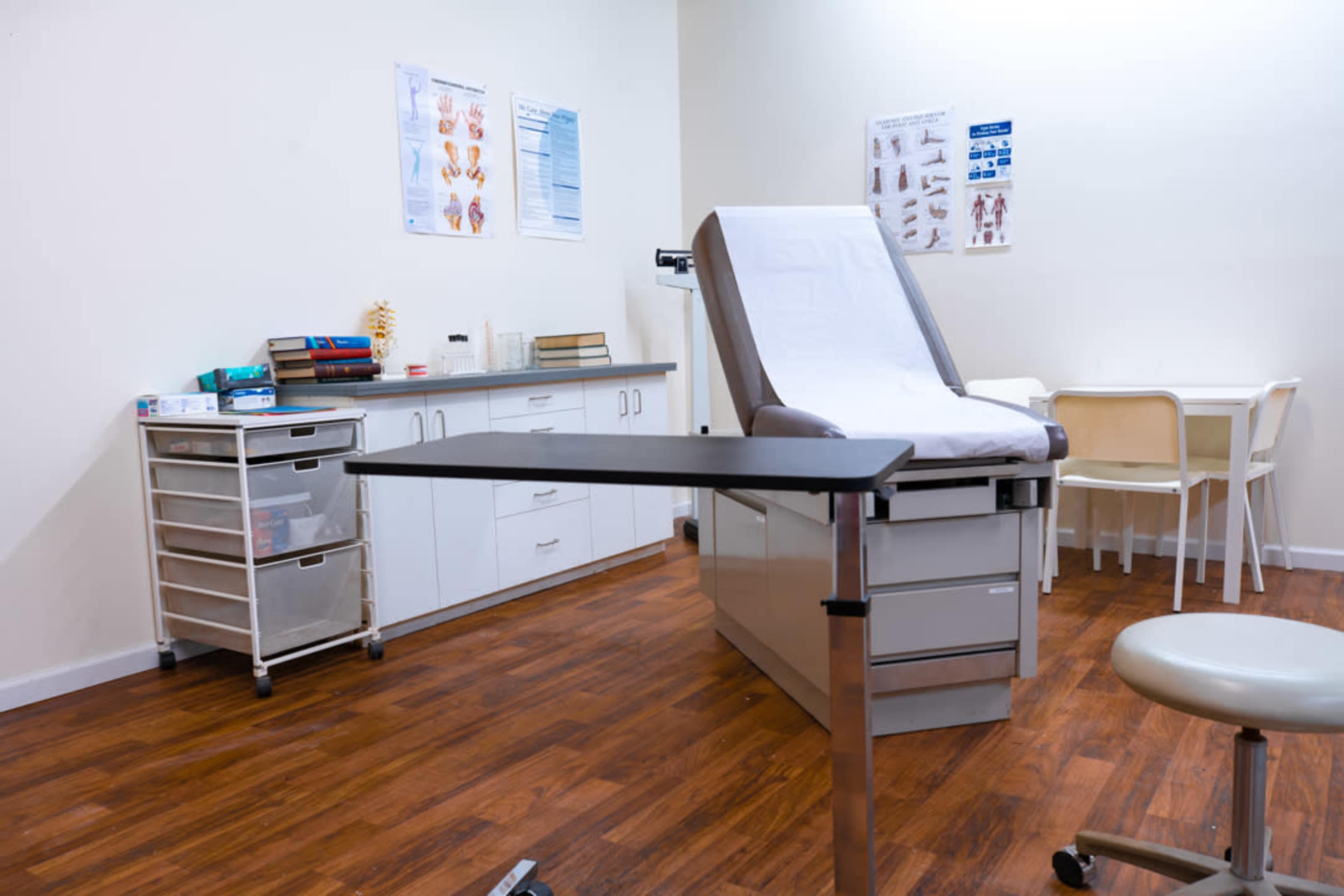 A medical examination room features an examination table, a cart with supplies, and a small table with chairs.