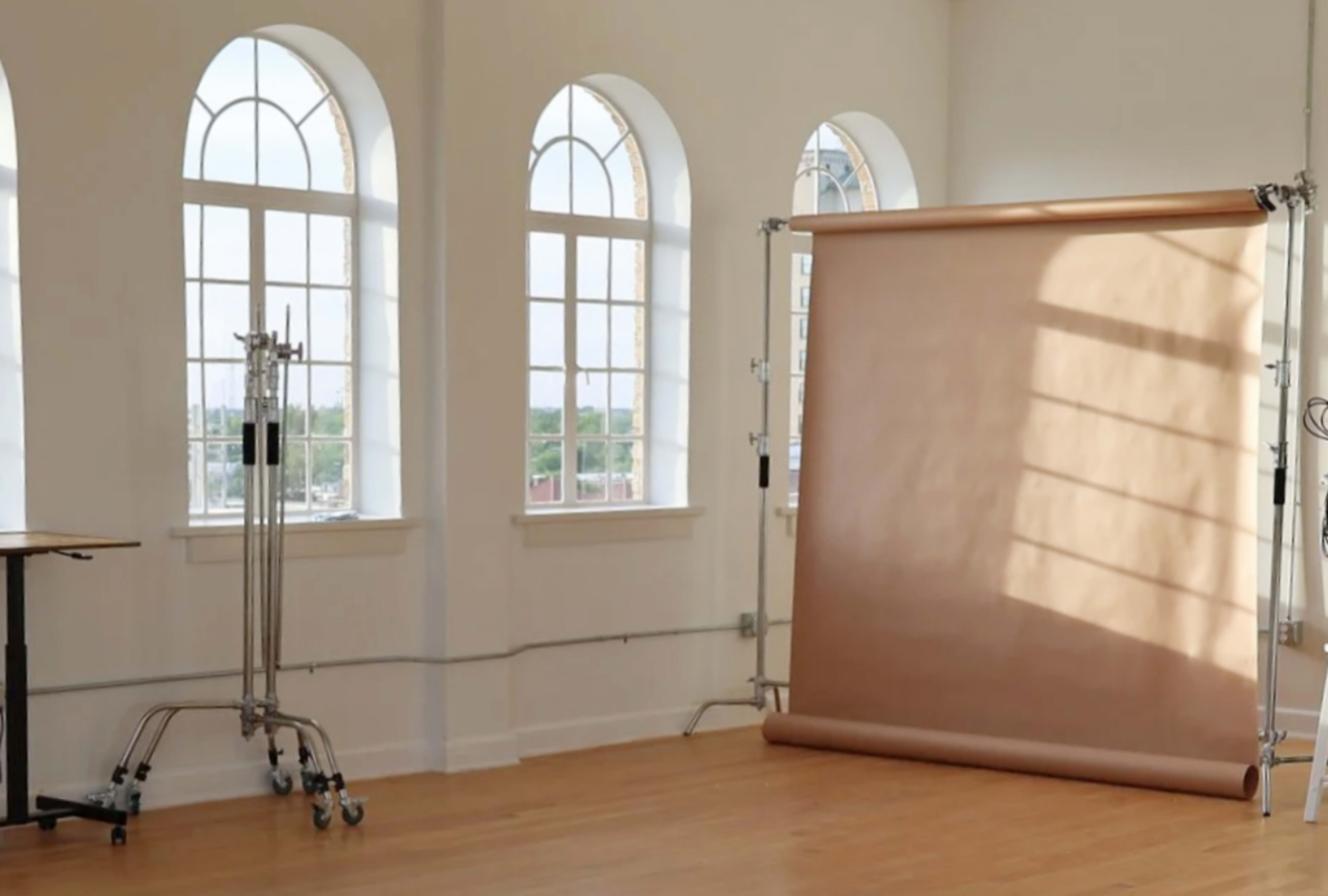 The image shows a well-lit studio space with arched windows, a backdrop stand with a rolled paper backdrop, and a table in the corner.