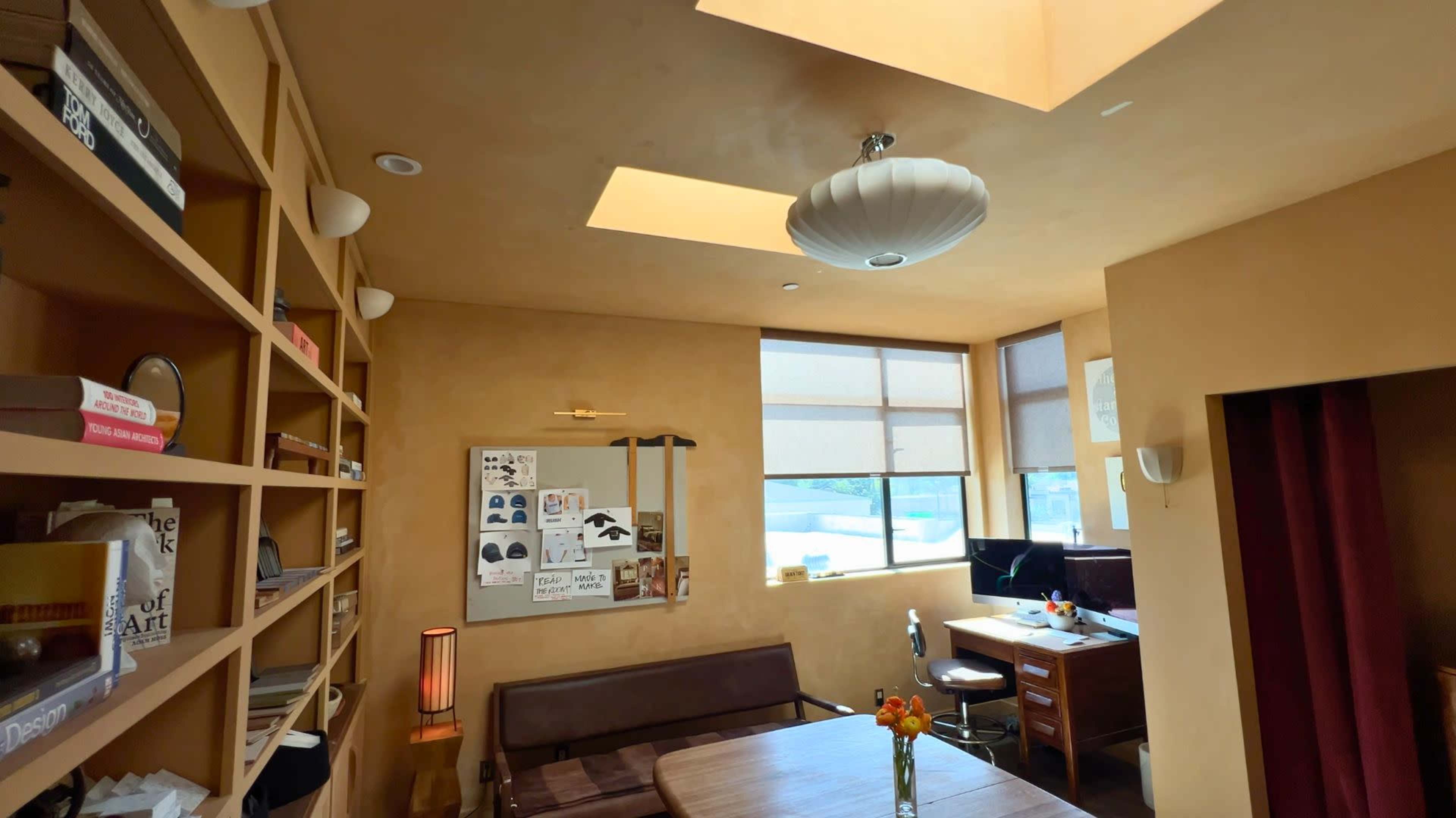 A cozy room features a wooden table, a seating area, a desk with computer monitors, and natural light coming through skylights and windows.