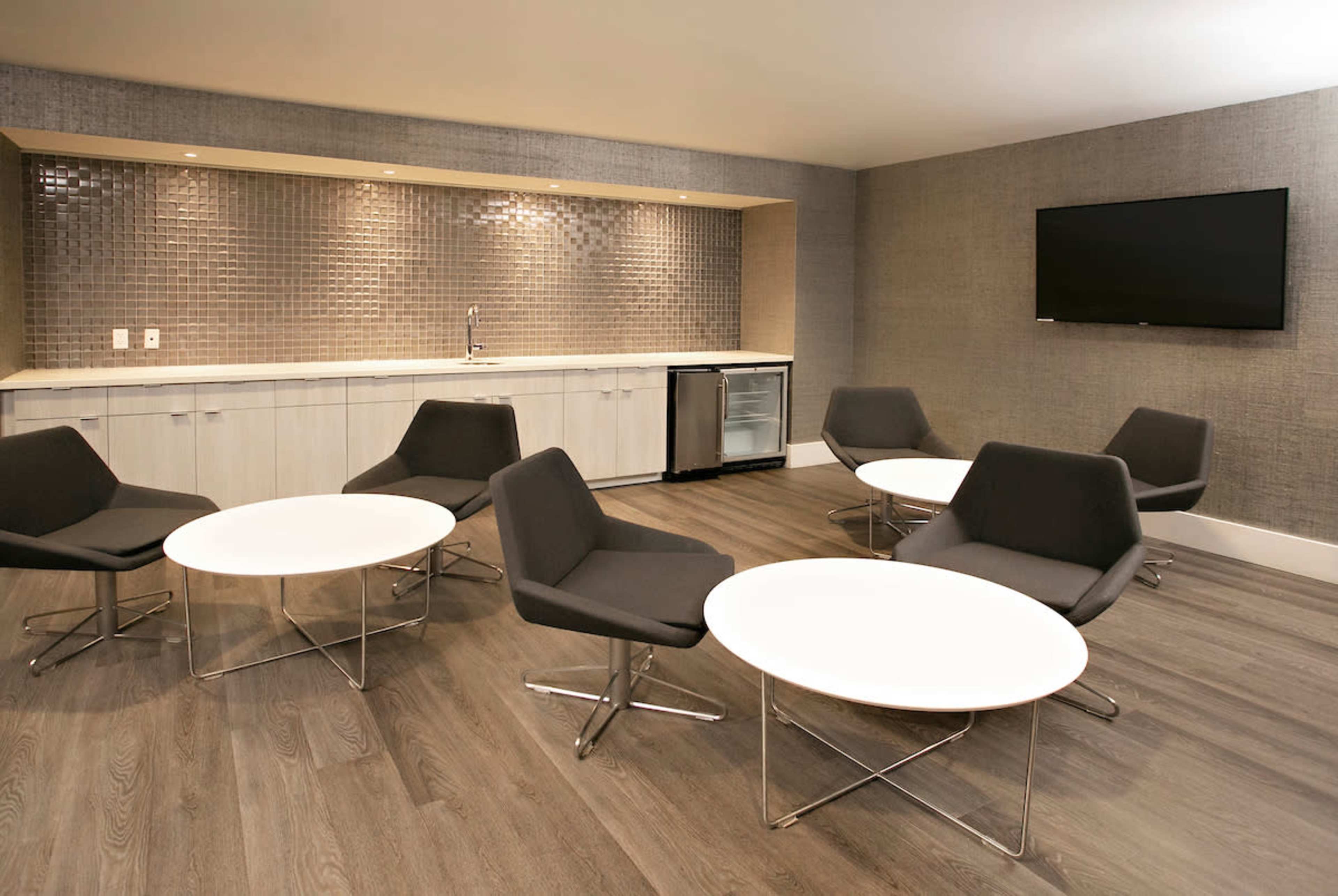 A modern lounge area features several gray chairs and round white tables, with a kitchenette and a television mounted on the wall.