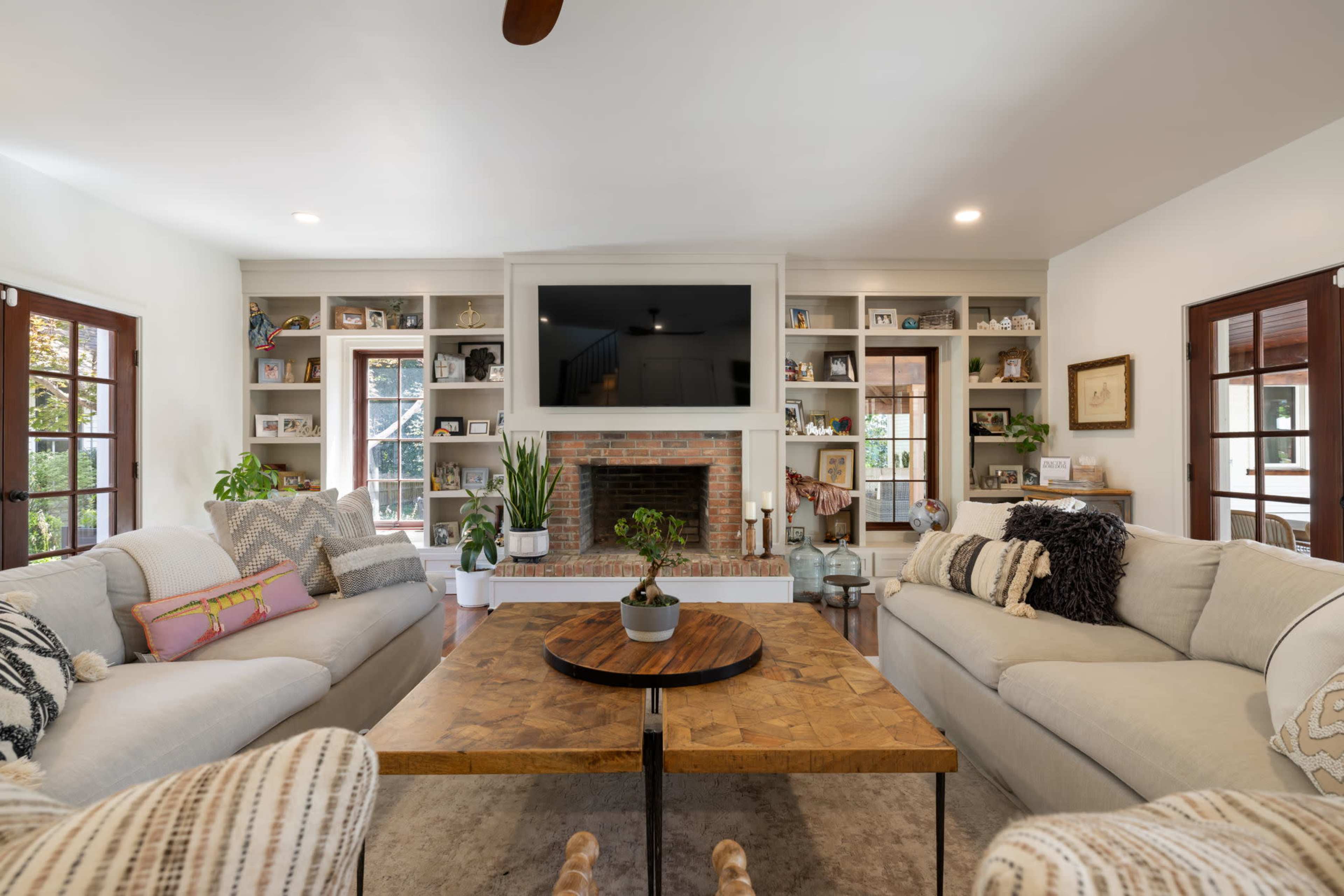 A spacious living room features two sofas facing a central wooden coffee table, a brick fireplace, and built-in shelves filled with decor.