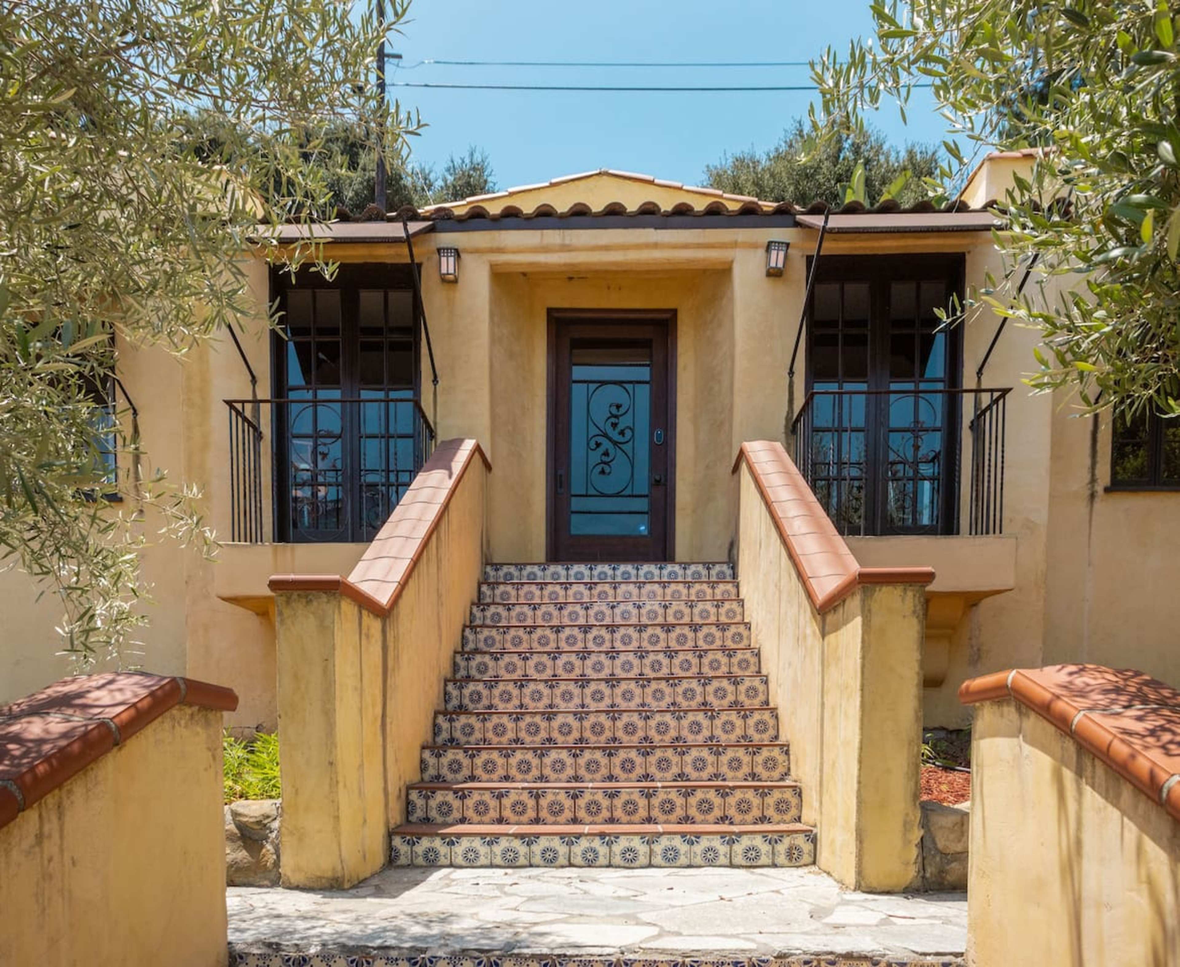 A stone pathway leads to a house with a decorative front door, flanked by large windows and a tiled staircase.