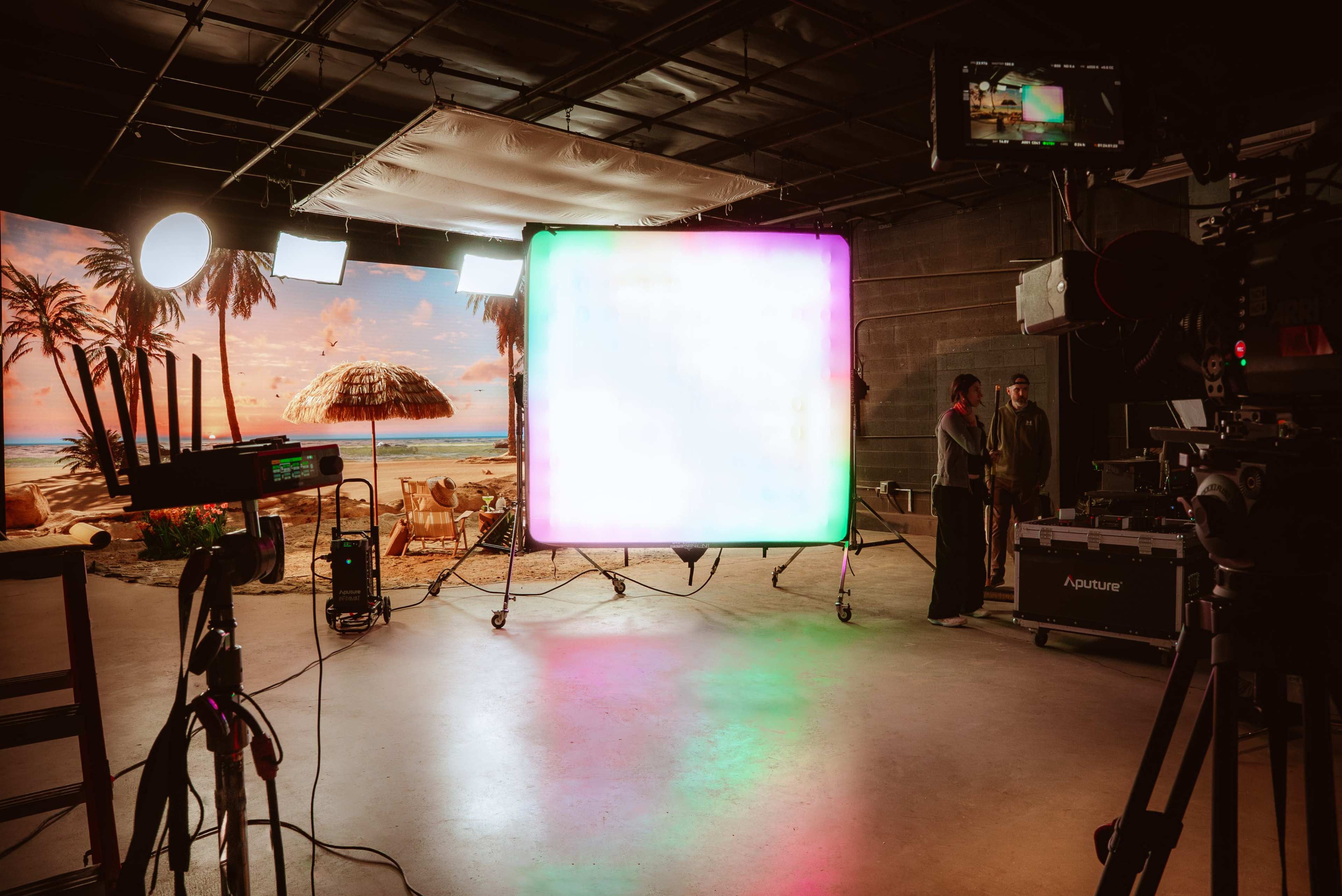 A film set features a brightly lit background of a tropical beach scene, with equipment and two crew members standing nearby.