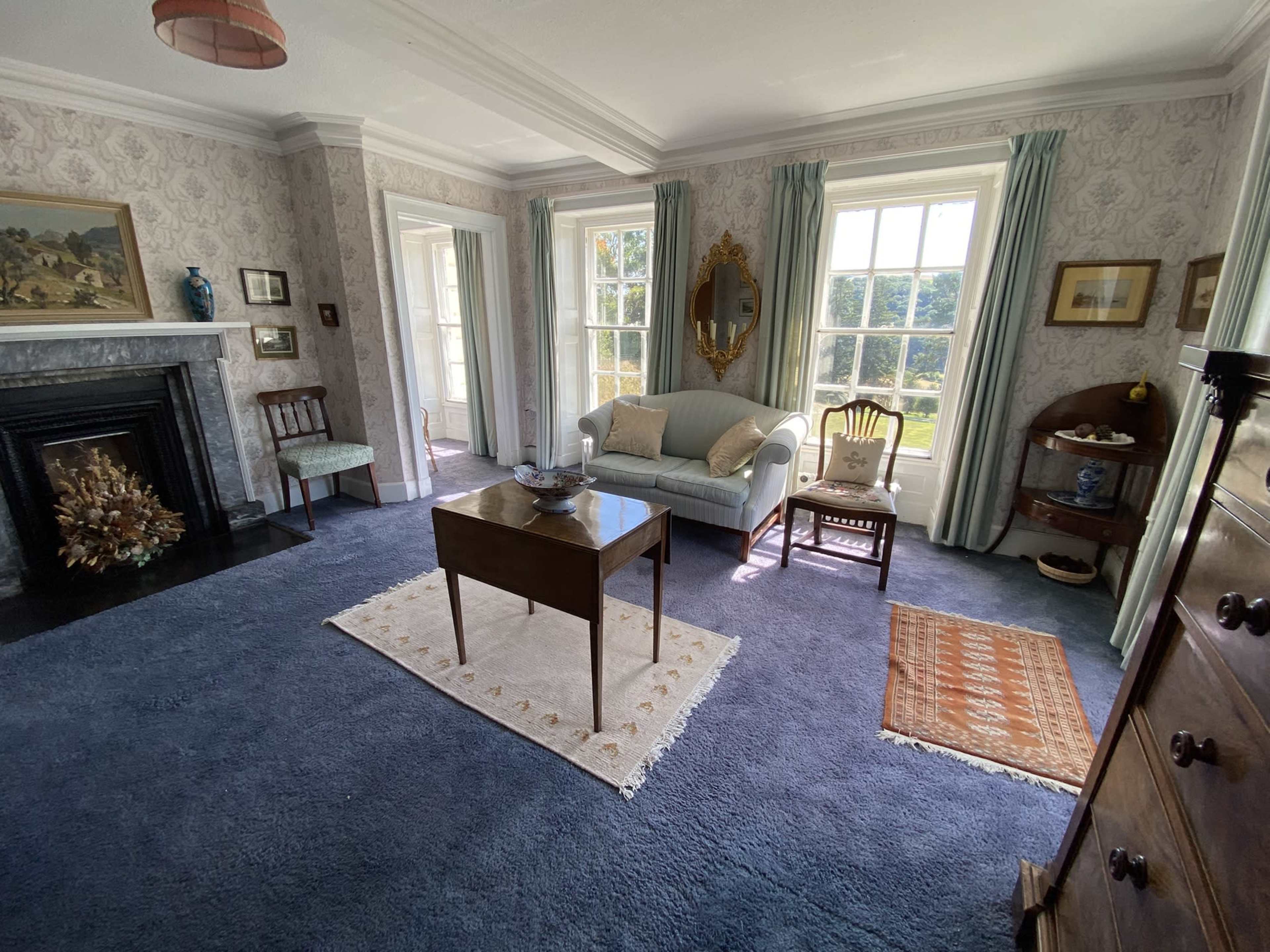 The image depicts a well-furnished living room with patterned wallpaper, a blue carpet, a wooden table in the center, and multiple windows allowing natural light to enter.