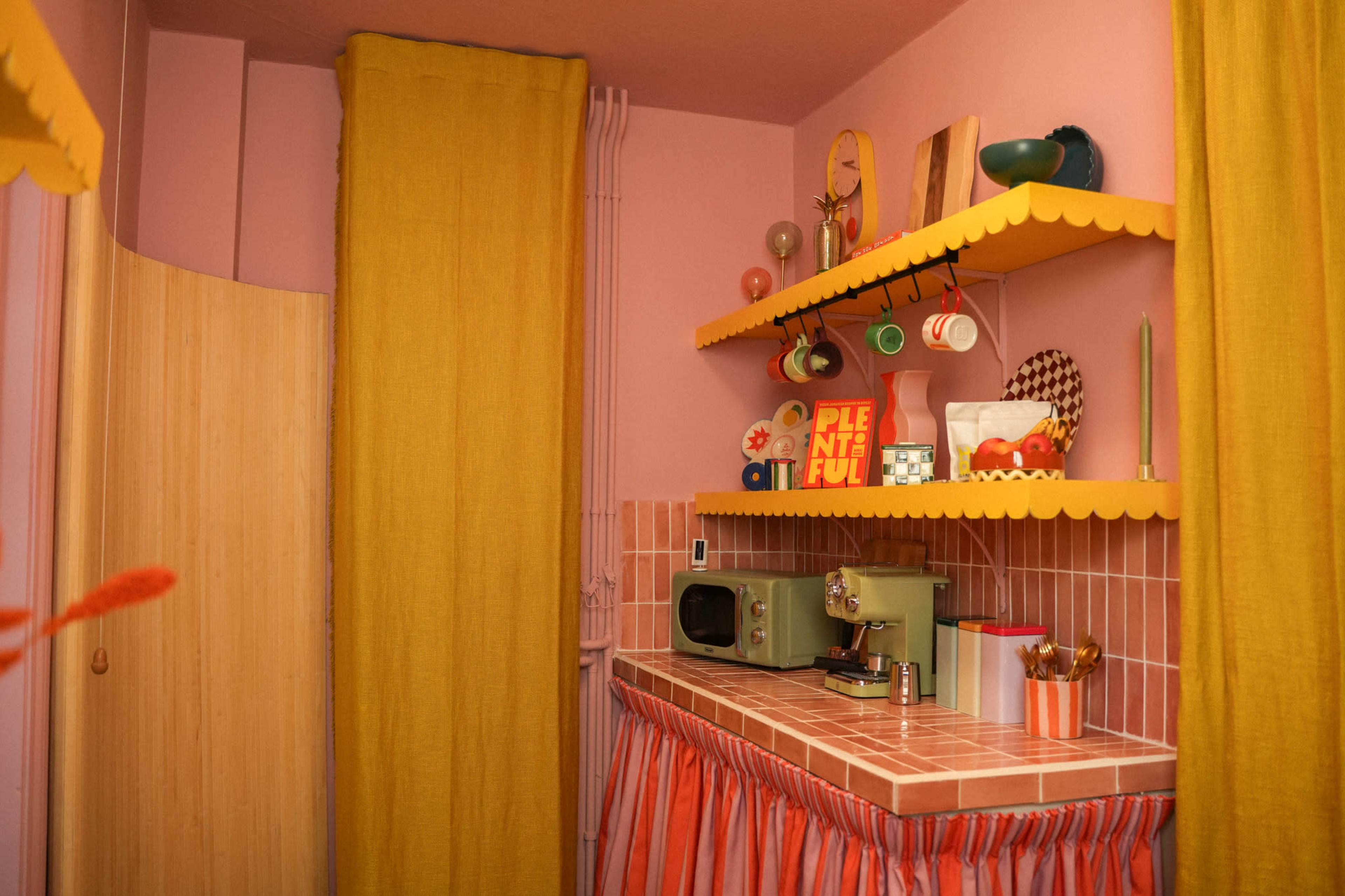 The image features a pastel-colored kitchen with pink walls, yellow curtains, and decorative shelves displaying colorful kitchen items.