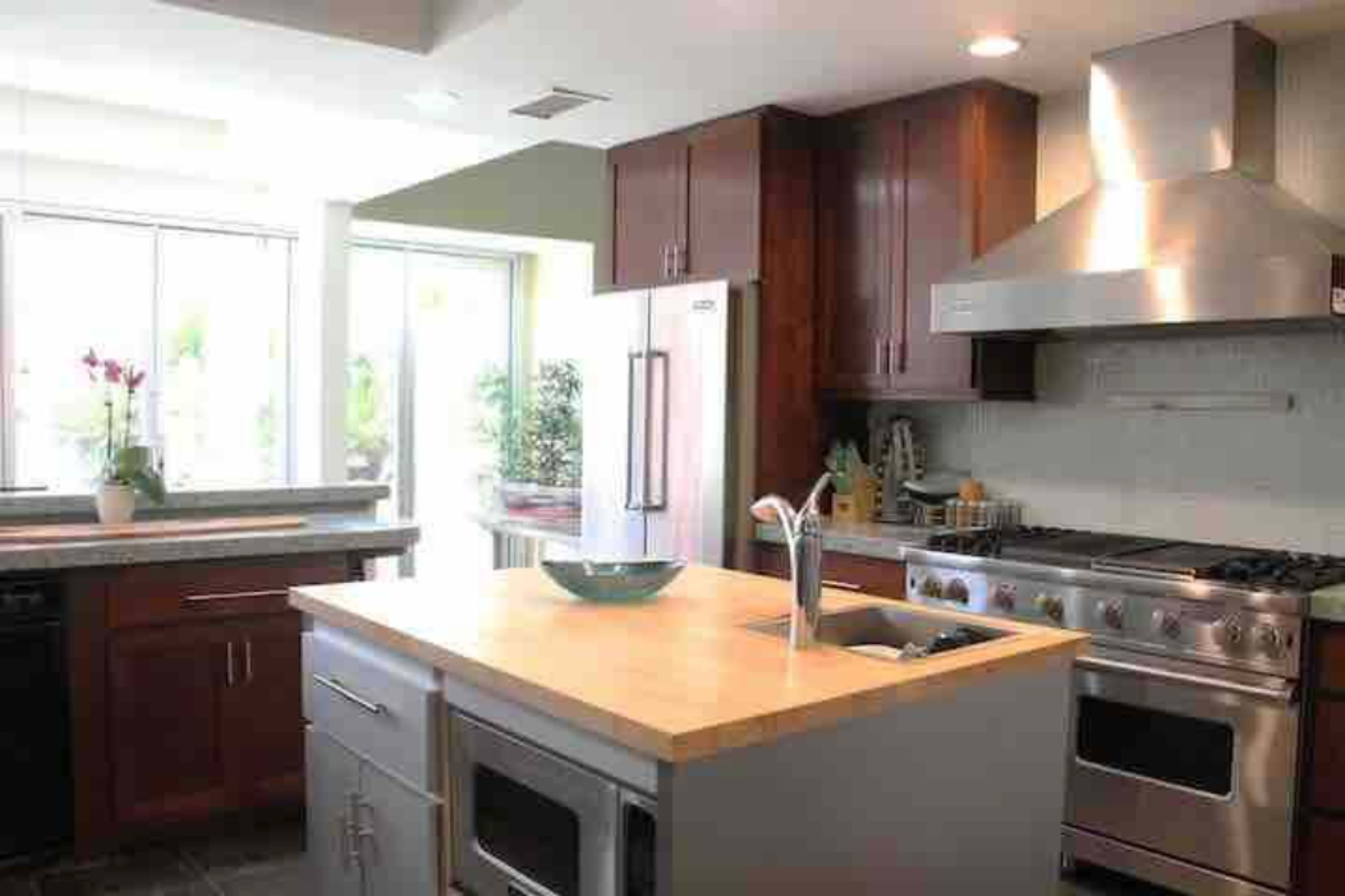 A modern kitchen with wooden cabinets, a large island featuring a wooden countertop, and a stainless steel range hood above a gas stove.