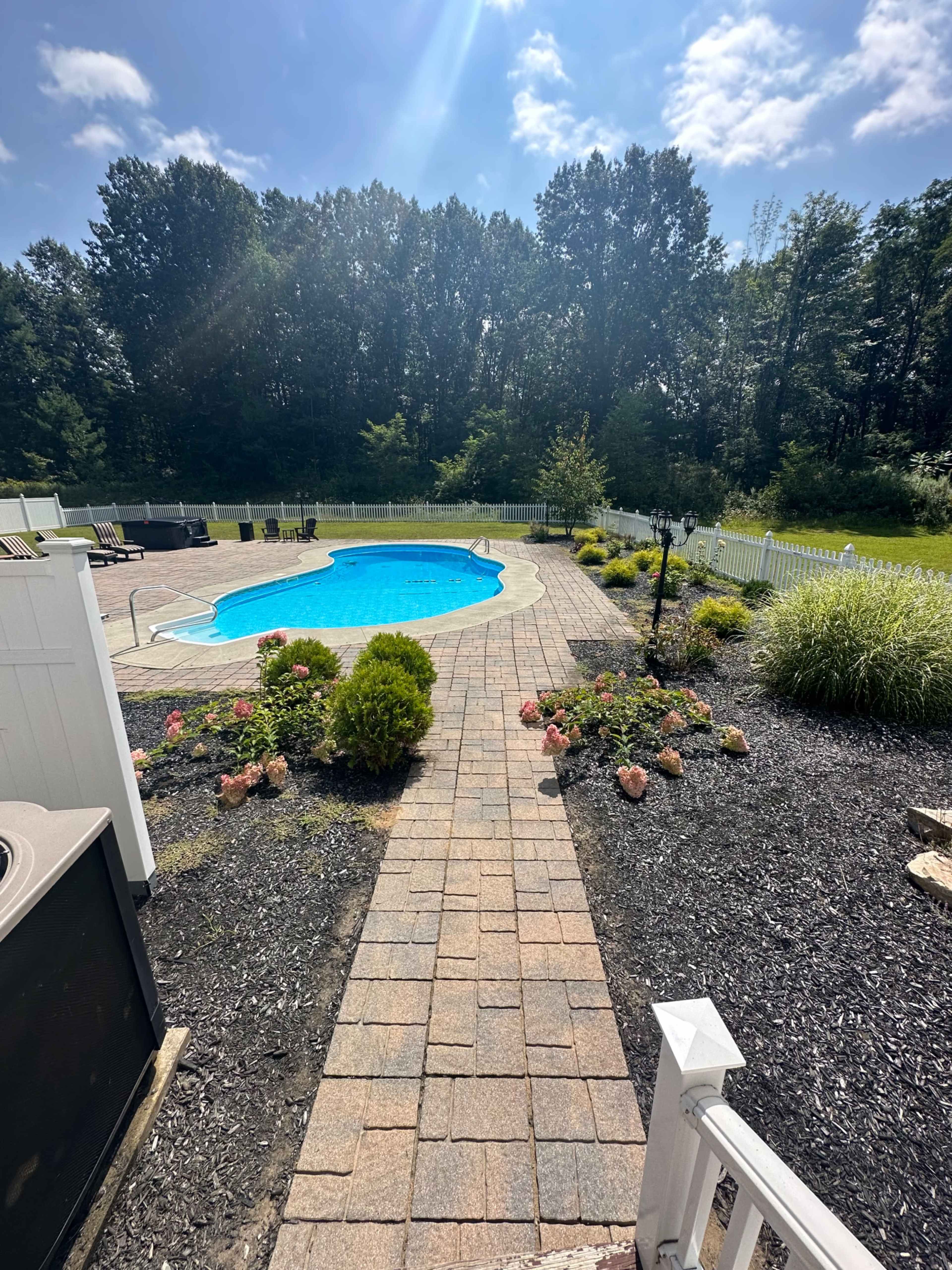 A stone pathway leads to a blue swimming pool surrounded by landscaped gardens and a wooded area in the background.
