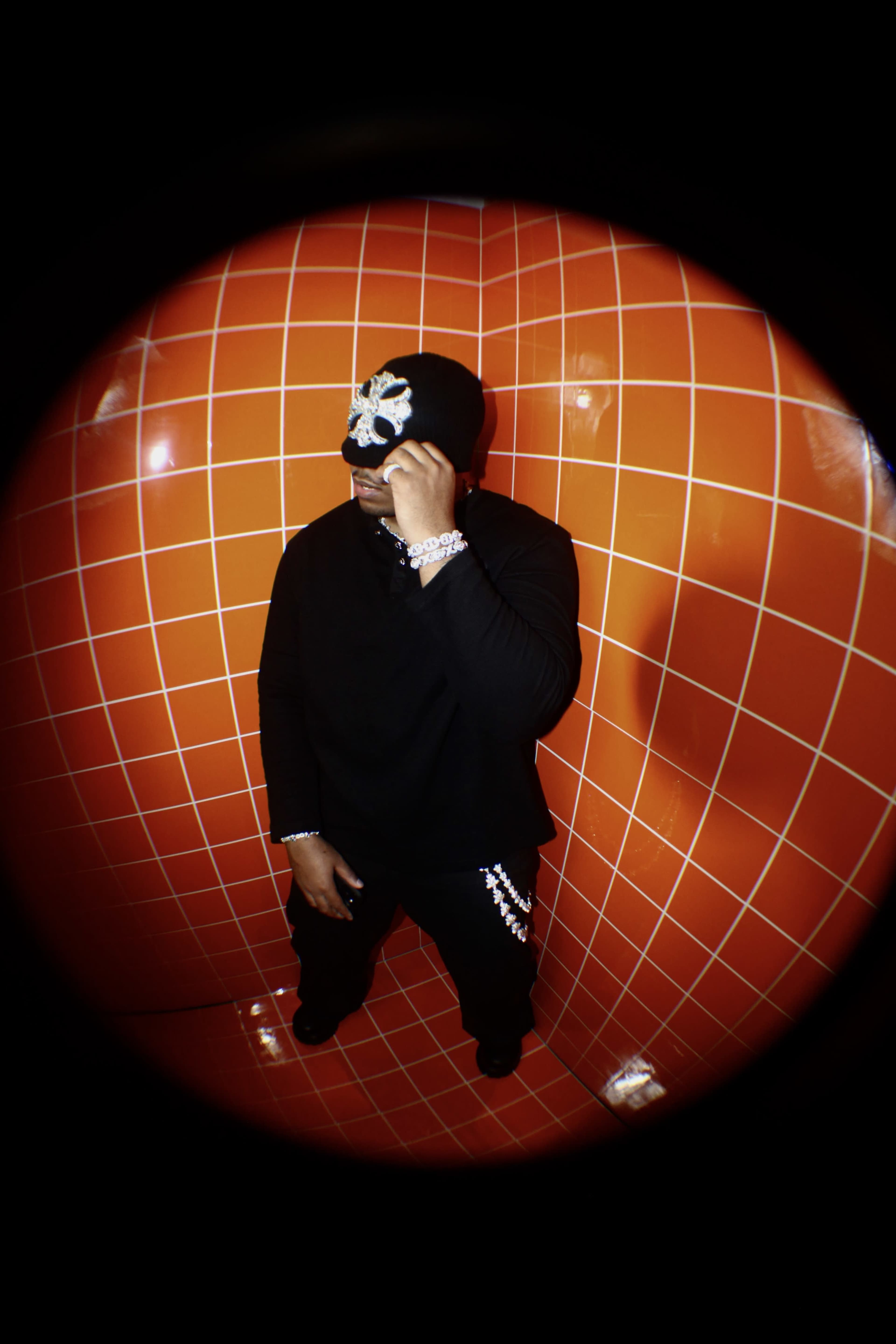 A person wearing a black outfit and a beanie stands in a corner with orange tiled walls.