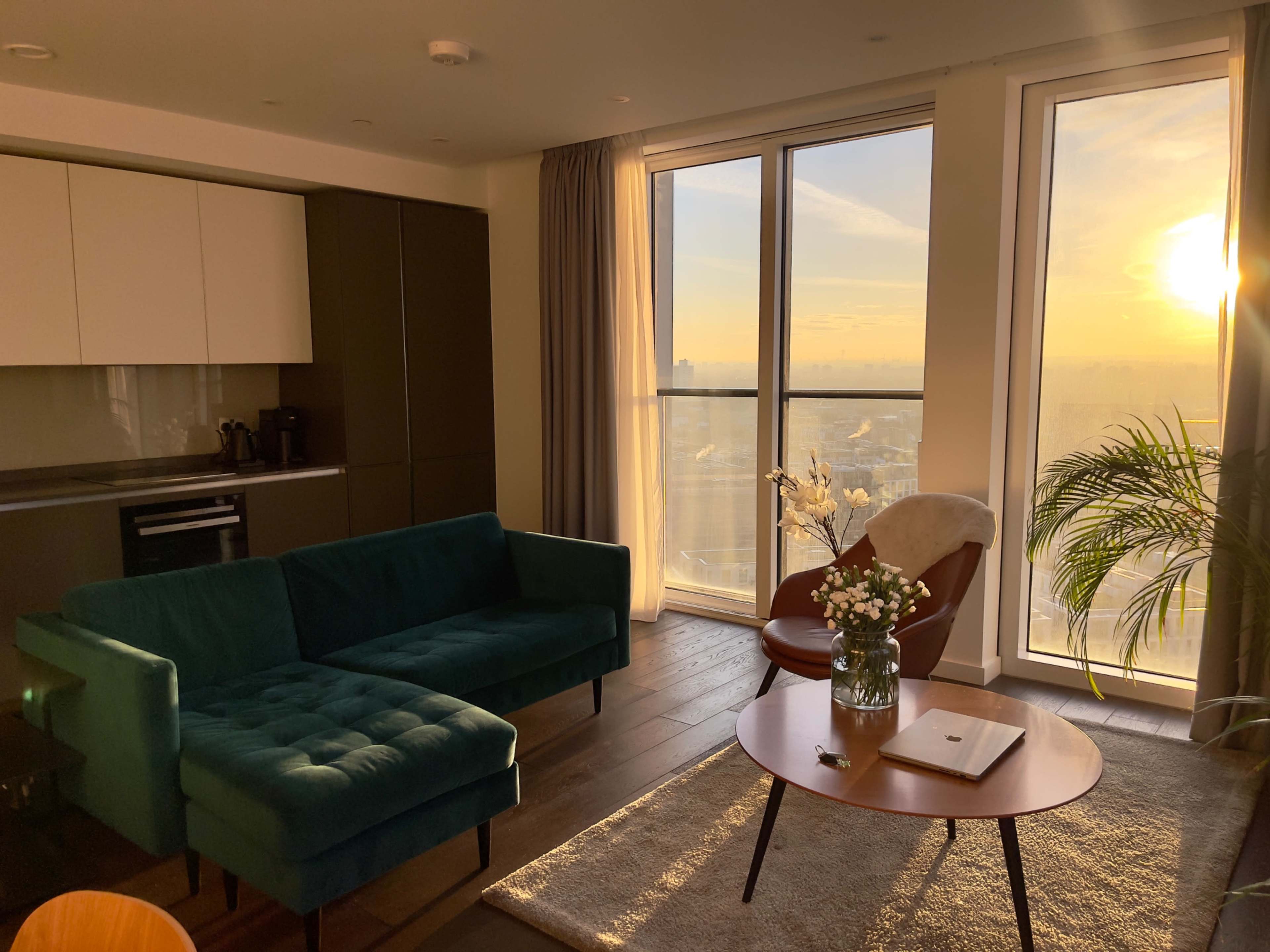 A bright living room features a teal sofa, a round table with a laptop, and large windows showcasing a sunset view over the city.