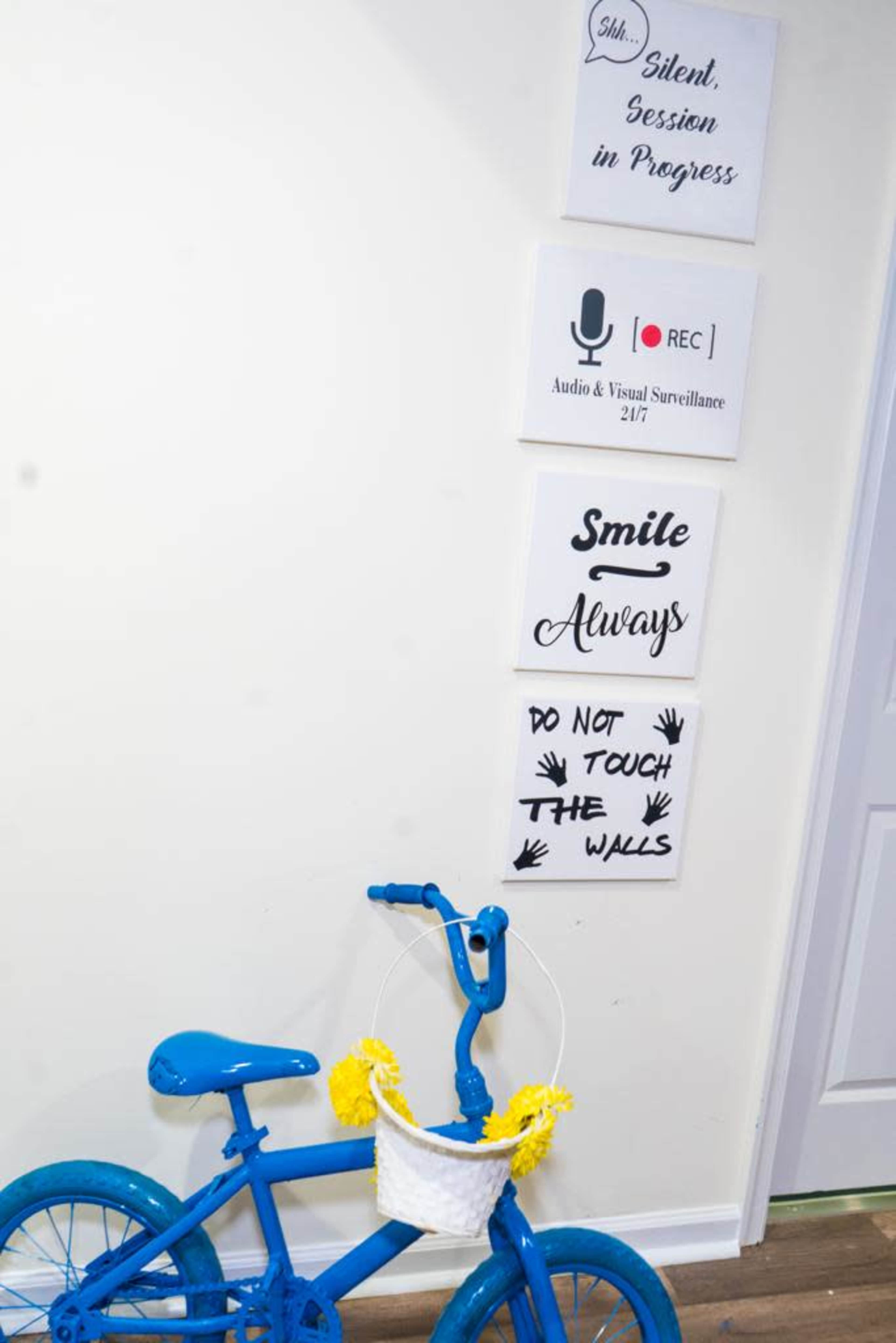 A blue bicycle with a decorative basket stands against a white wall adorned with various signs related to a silent session and visual surveillance.