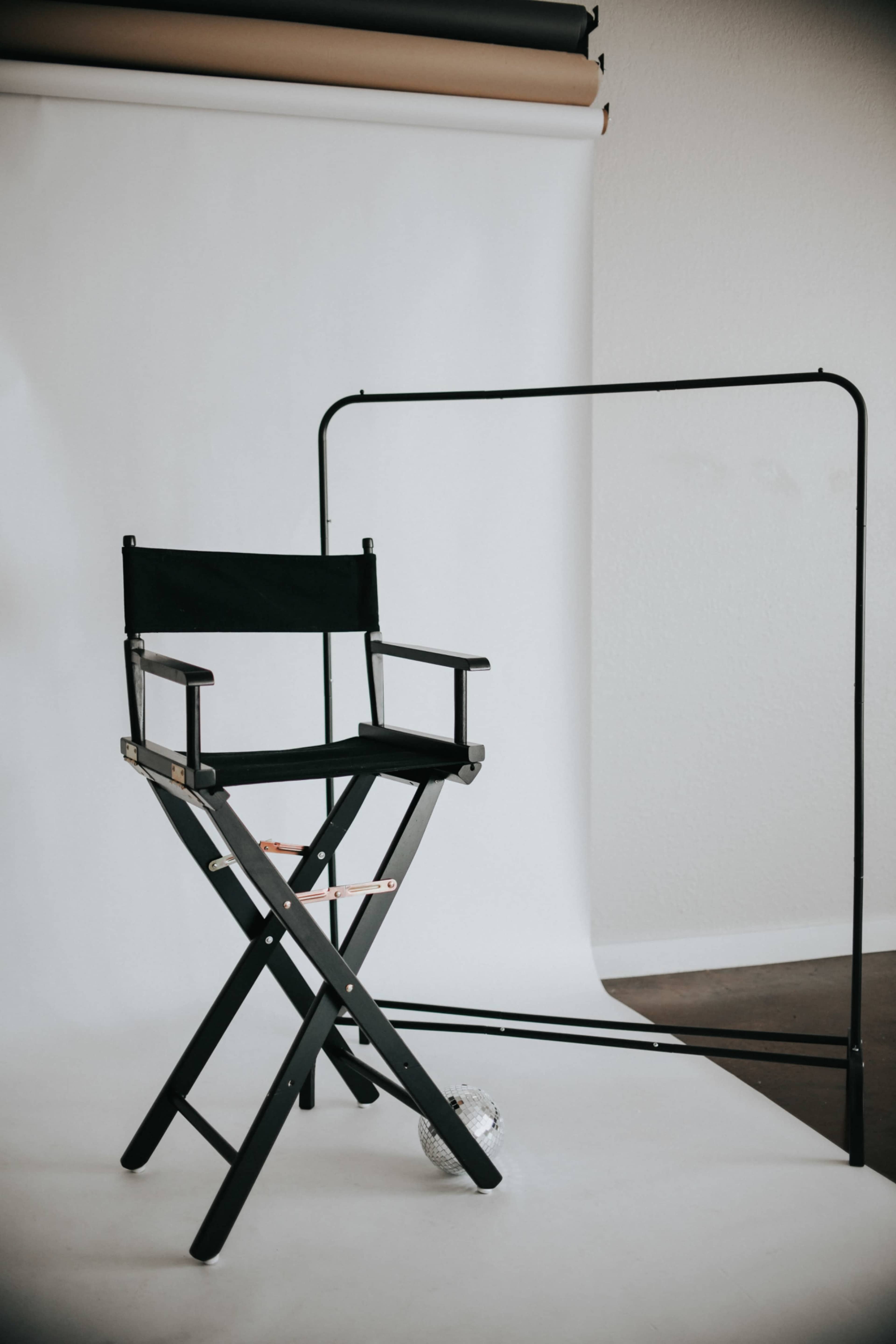 A black director's chair stands in front of a blank white backdrop, accompanied by a clothing rack and a small disco ball on the floor.