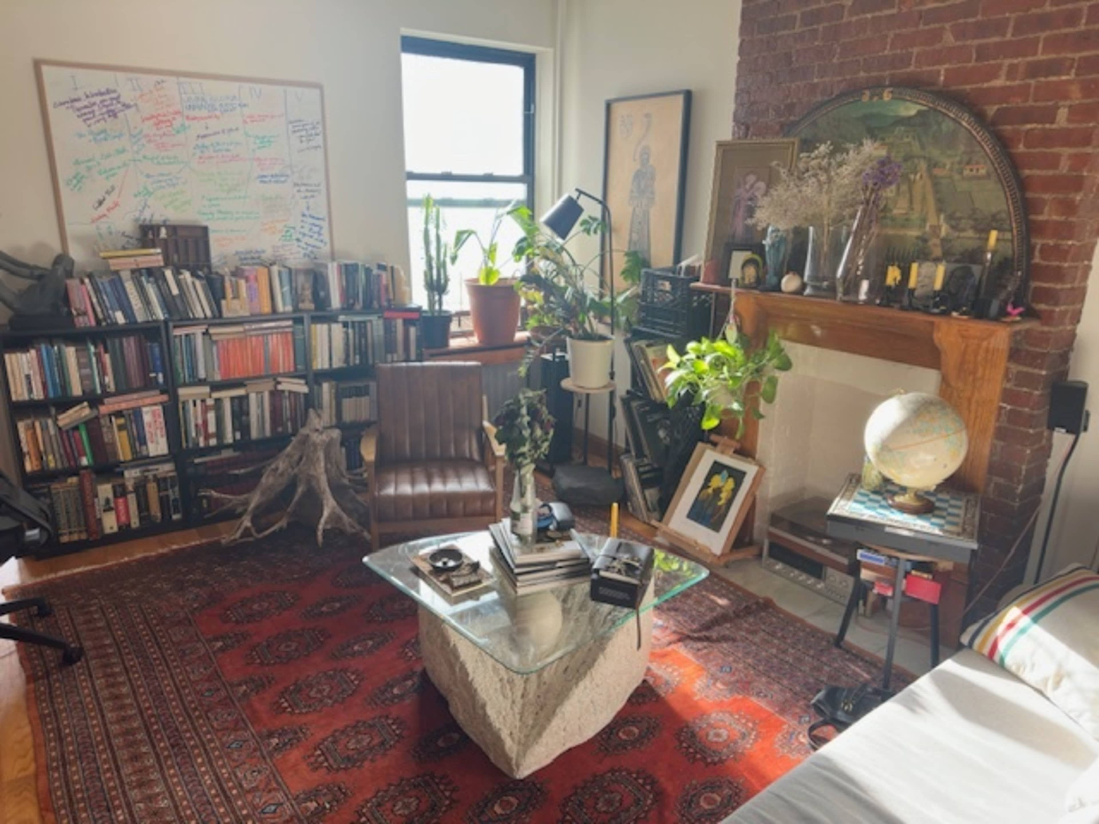 The image shows a cozy room with a bookshelf filled with books, a glass coffee table, a brick fireplace, and various plants and decorative items.