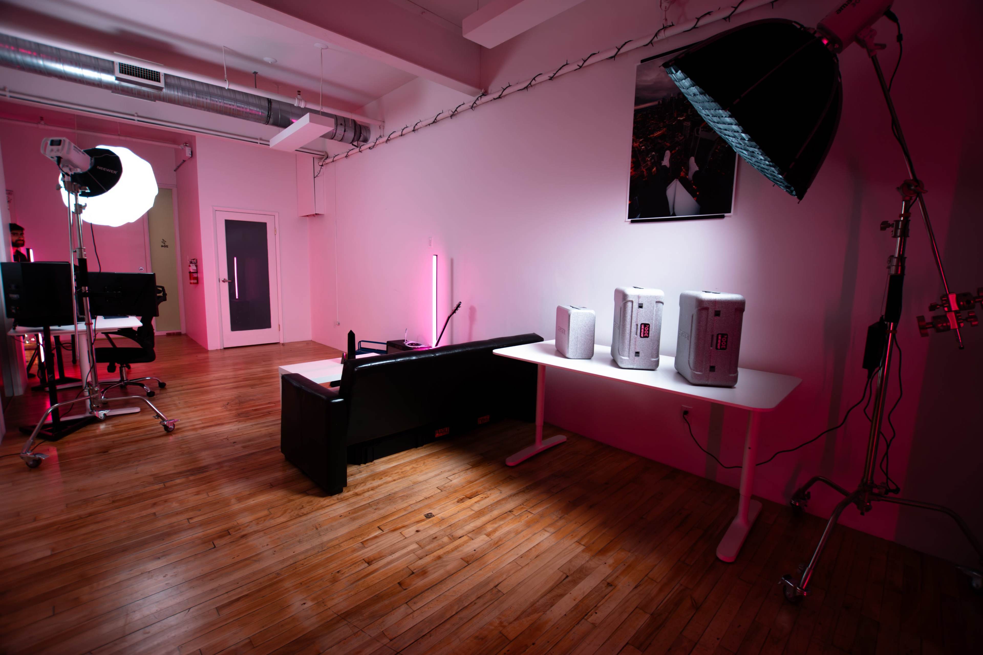 The image shows a modern studio space featuring a black couch, a white table with three speakers, and photography lighting equipment under soft pink lighting.