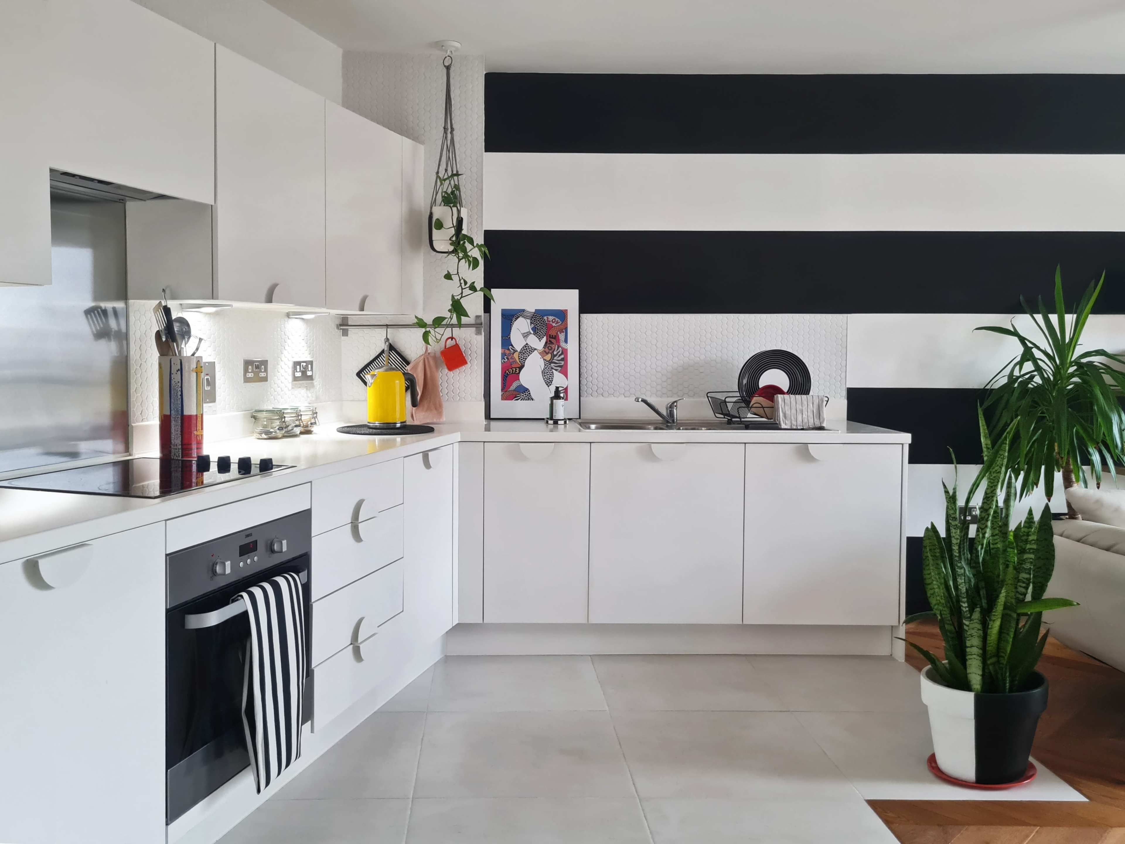 A modern kitchen with white cabinets, a black striped wall, and a yellow kettle on the counter beside a framed picture.