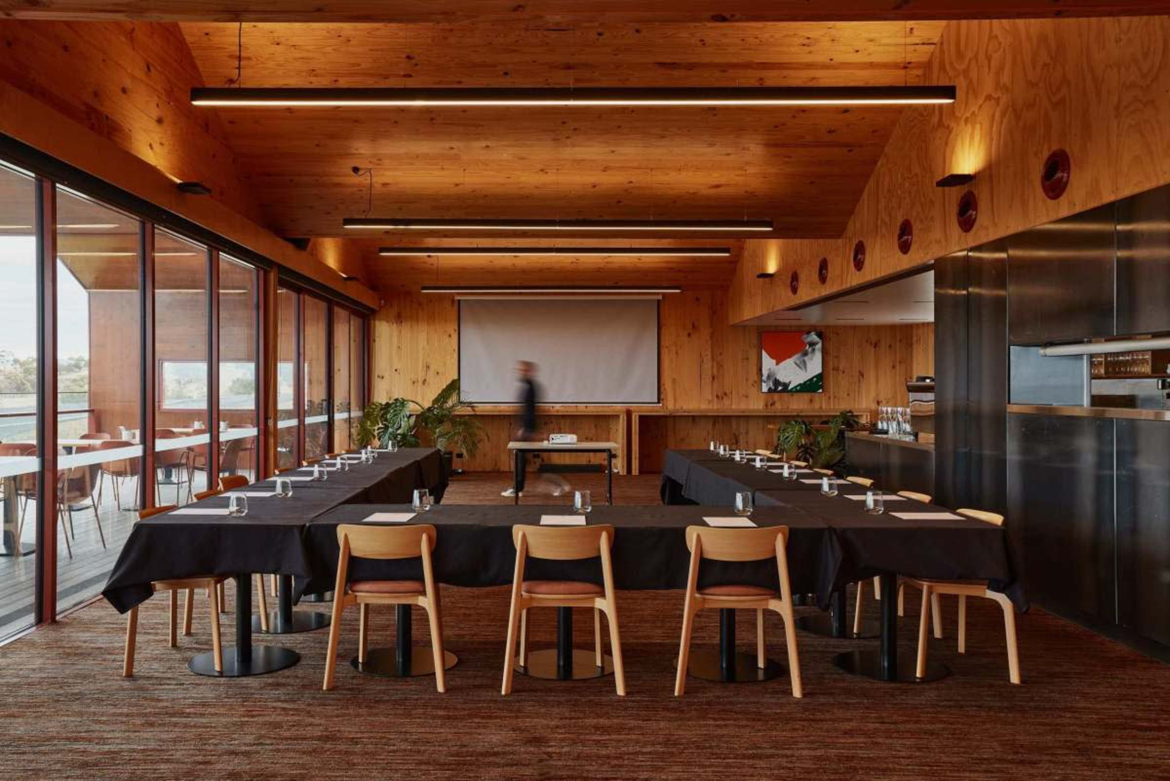 A spacious meeting room features a long table set with water glasses and napkins, surrounded by wooden chairs, with large windows providing a view outside.