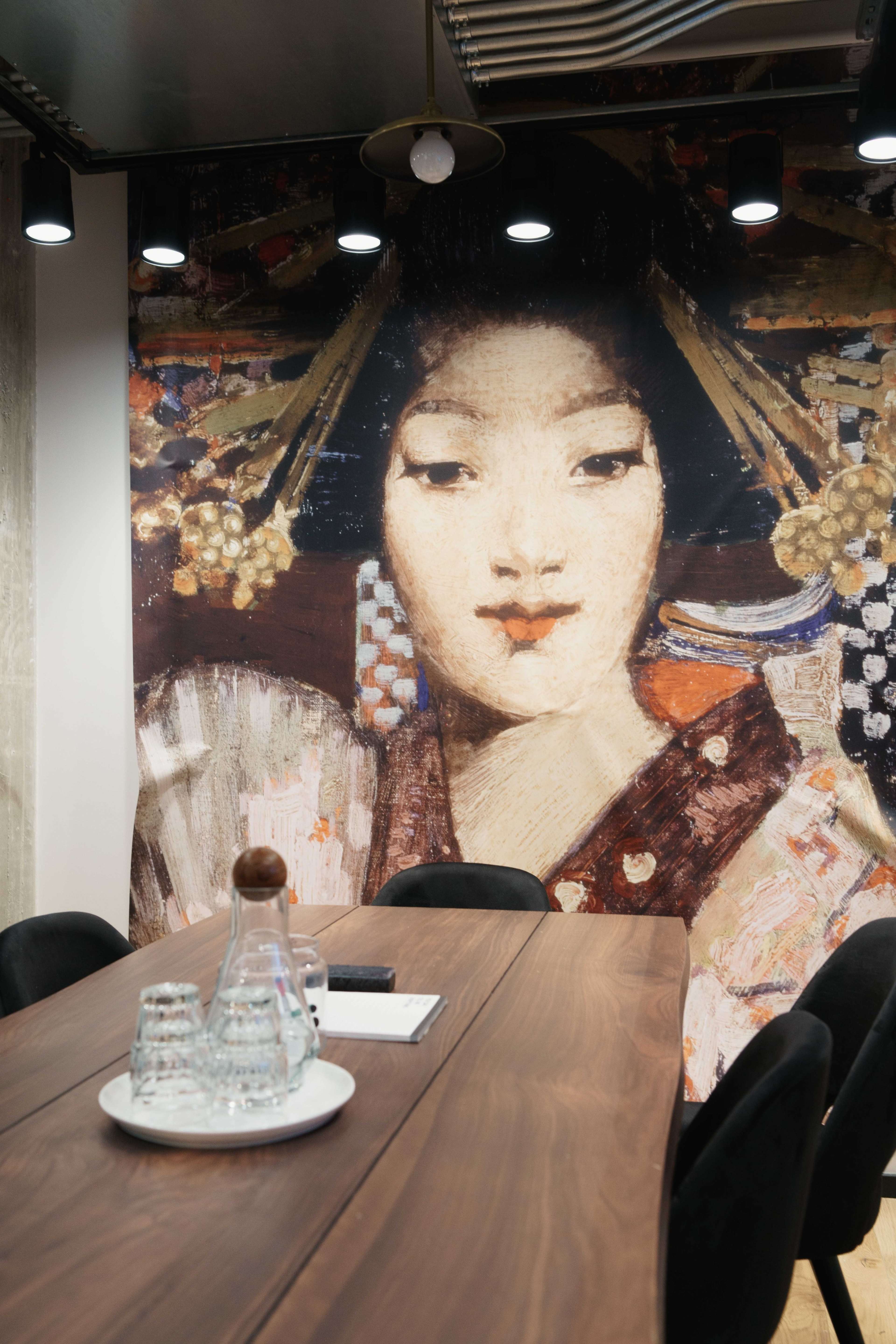 A wooden conference table is set with glassware and a notepad in front of a large mural depicting a traditional Japanese woman.