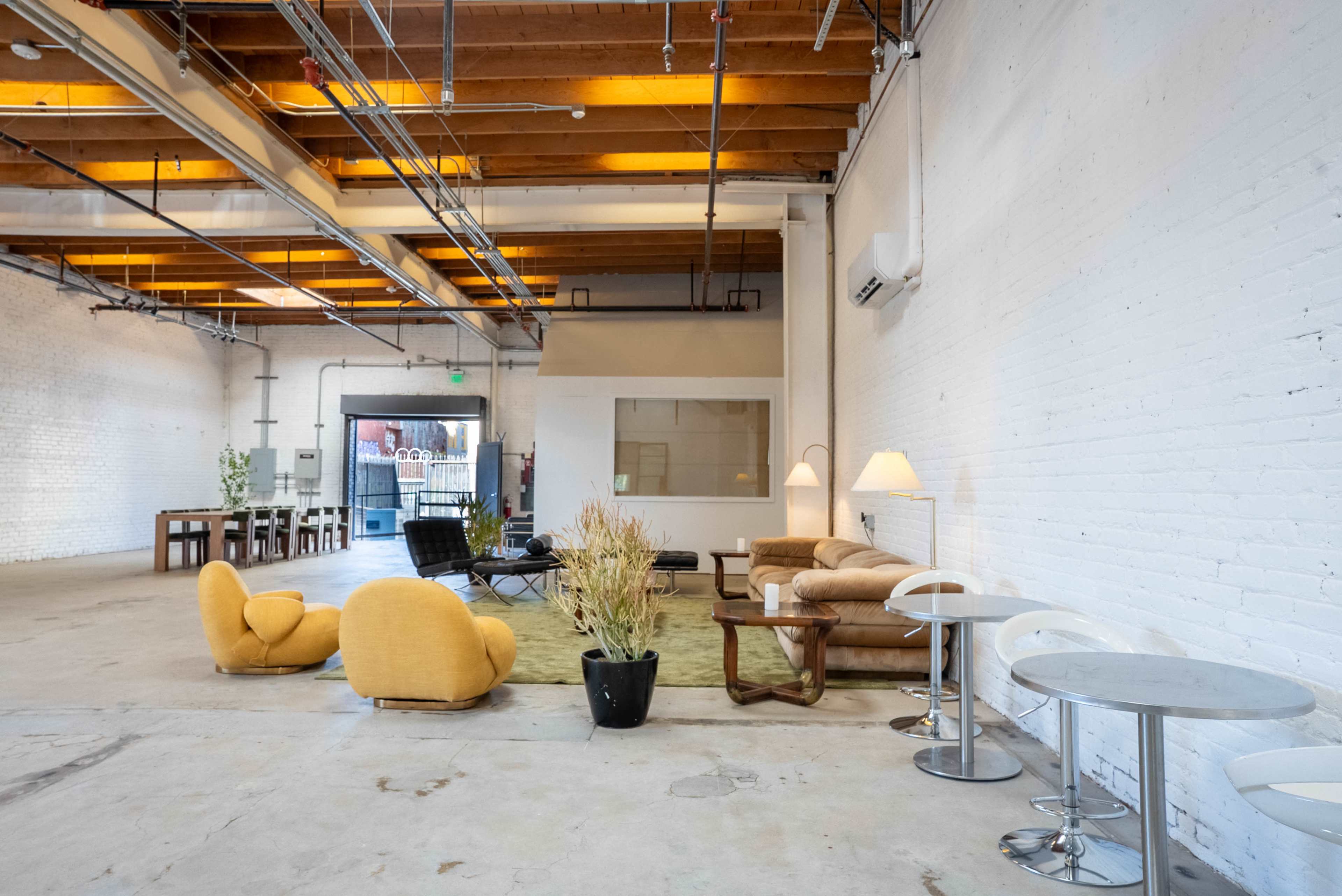 The image shows an open industrial-style interior space with a mix of seating areas, including a yellow lounge chair and sofas, and tables set up against the walls.