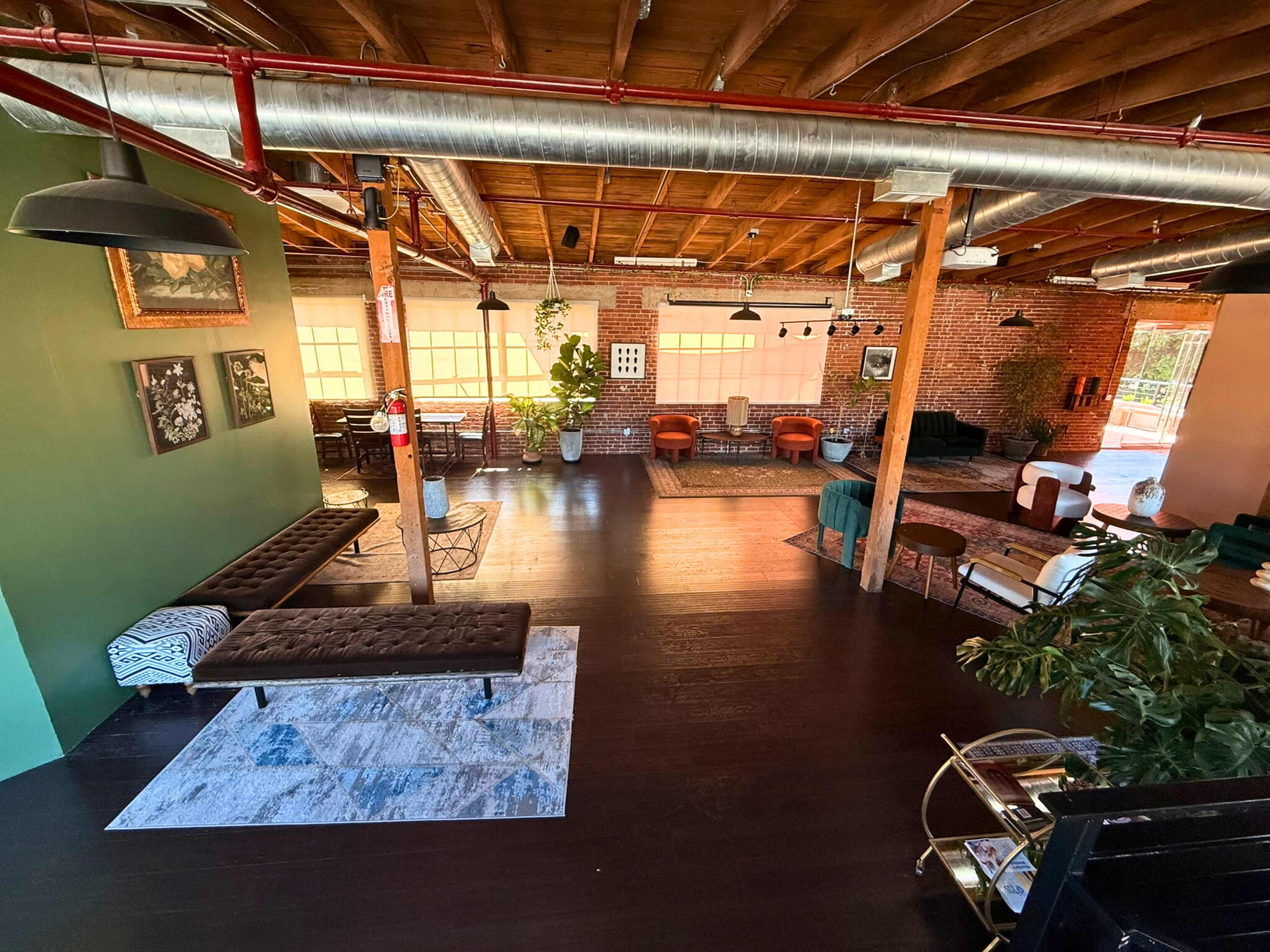 The image shows a spacious, modern interior with wooden beams and a mix of seating arrangements, including couches and chairs, surrounded by large windows and green walls.