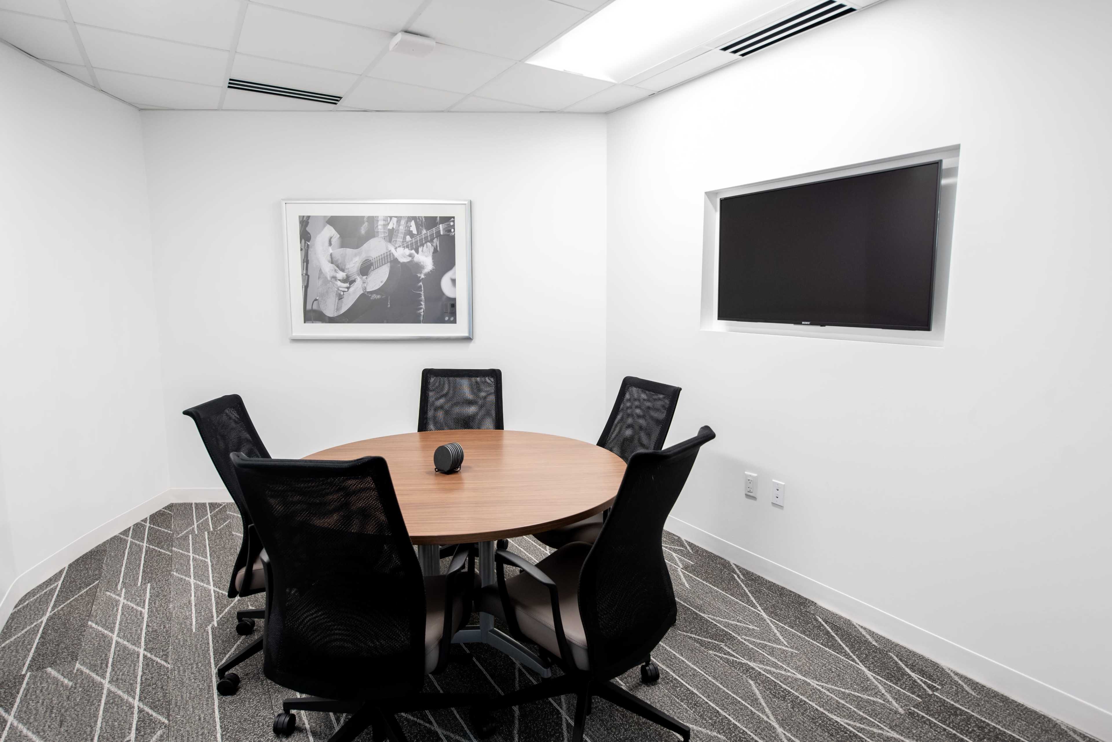 A small meeting room features a round wooden table surrounded by five black mesh chairs, with a framed photo on the wall and a mounted screen.