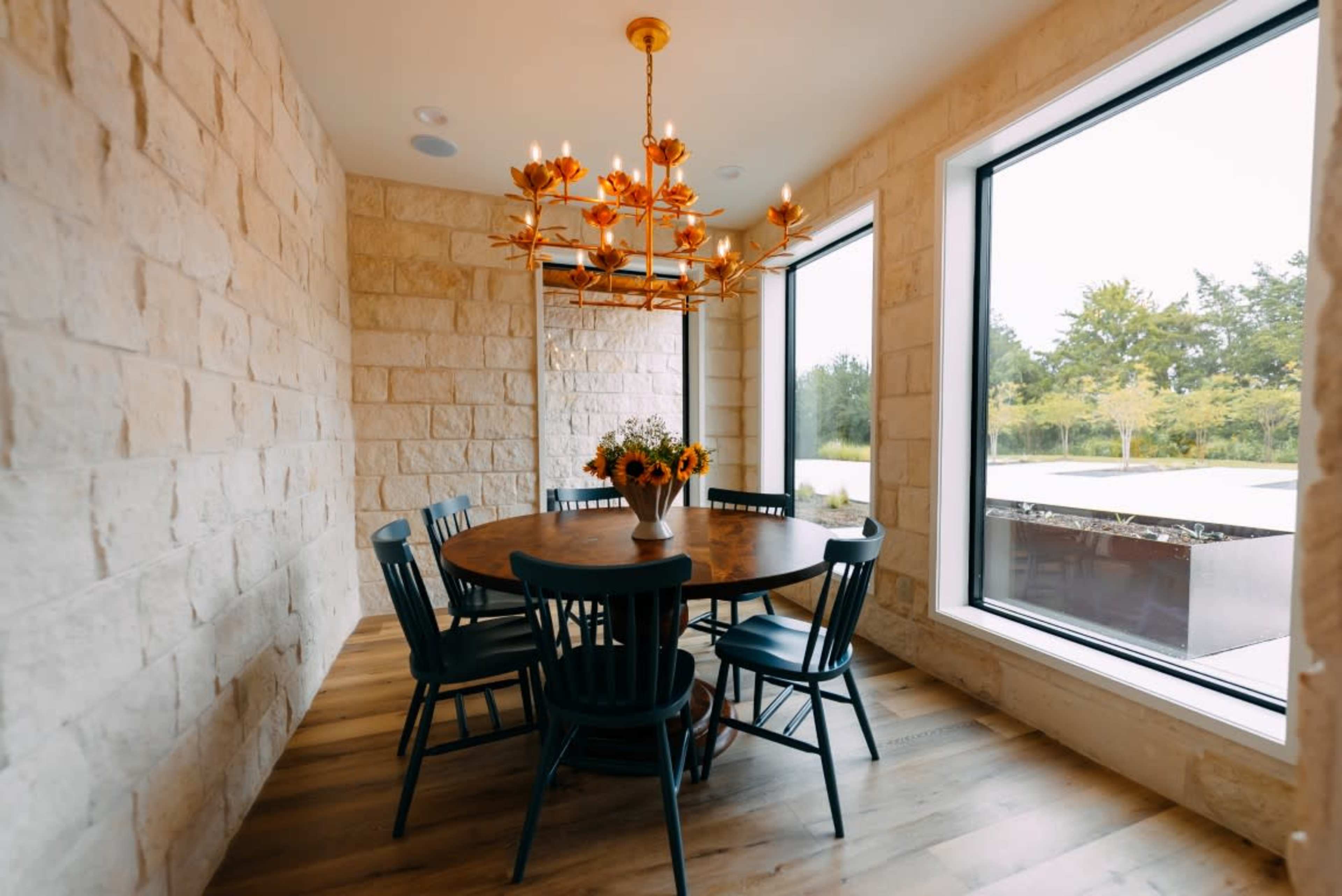 A circular wooden dining table surrounded by six dark chairs is illuminated by a large chandelier in a room with stone walls and large windows.