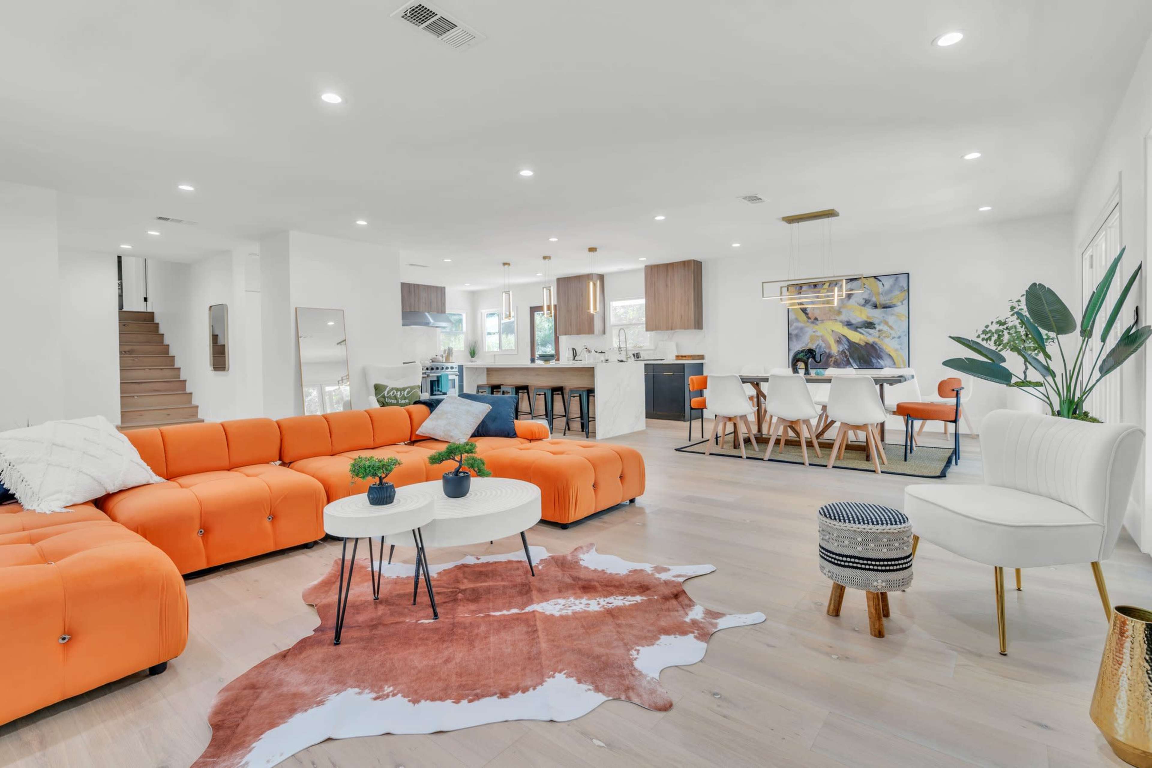 A modern living area features an orange sectional sofa, a round coffee table, and a dining space with a wooden table, surrounded by white walls and wooden flooring.