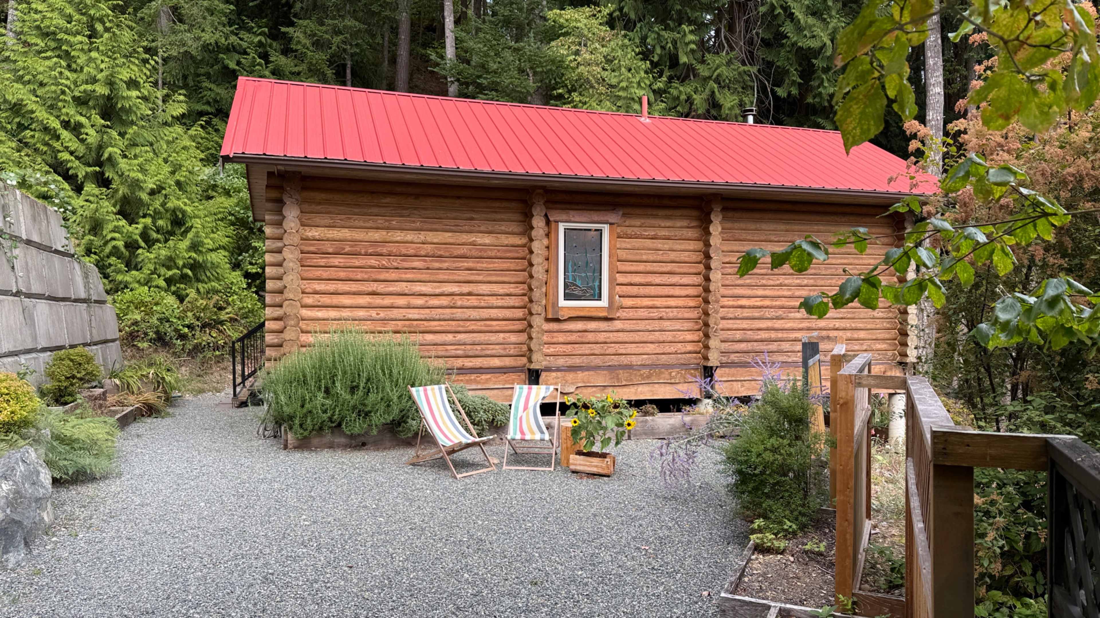 The image shows a log cabin with a red metal roof, surrounded by trees and a gravel walkway, featuring two striped lounge chairs and a flower pot in front.