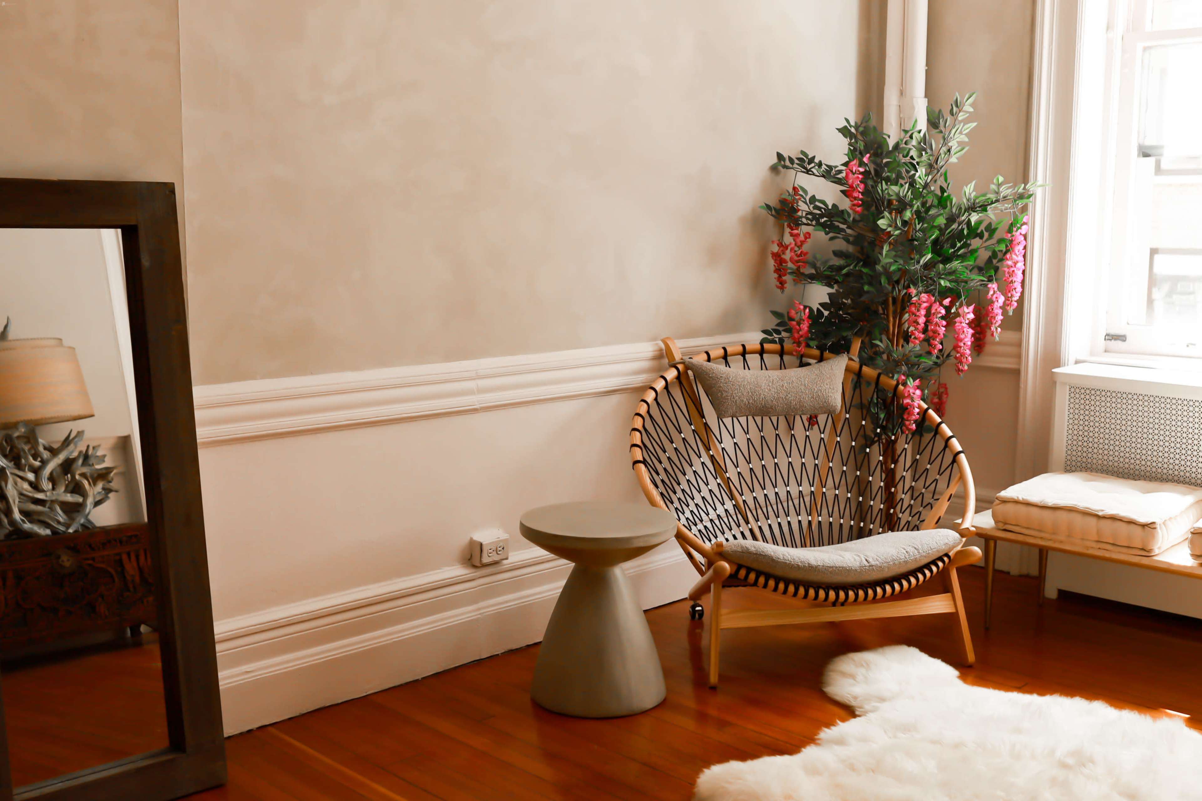 A cozy corner of a room features a wicker chair with a cushion, a small side table, a potted plant, and a fluffy rug on a wooden floor.
