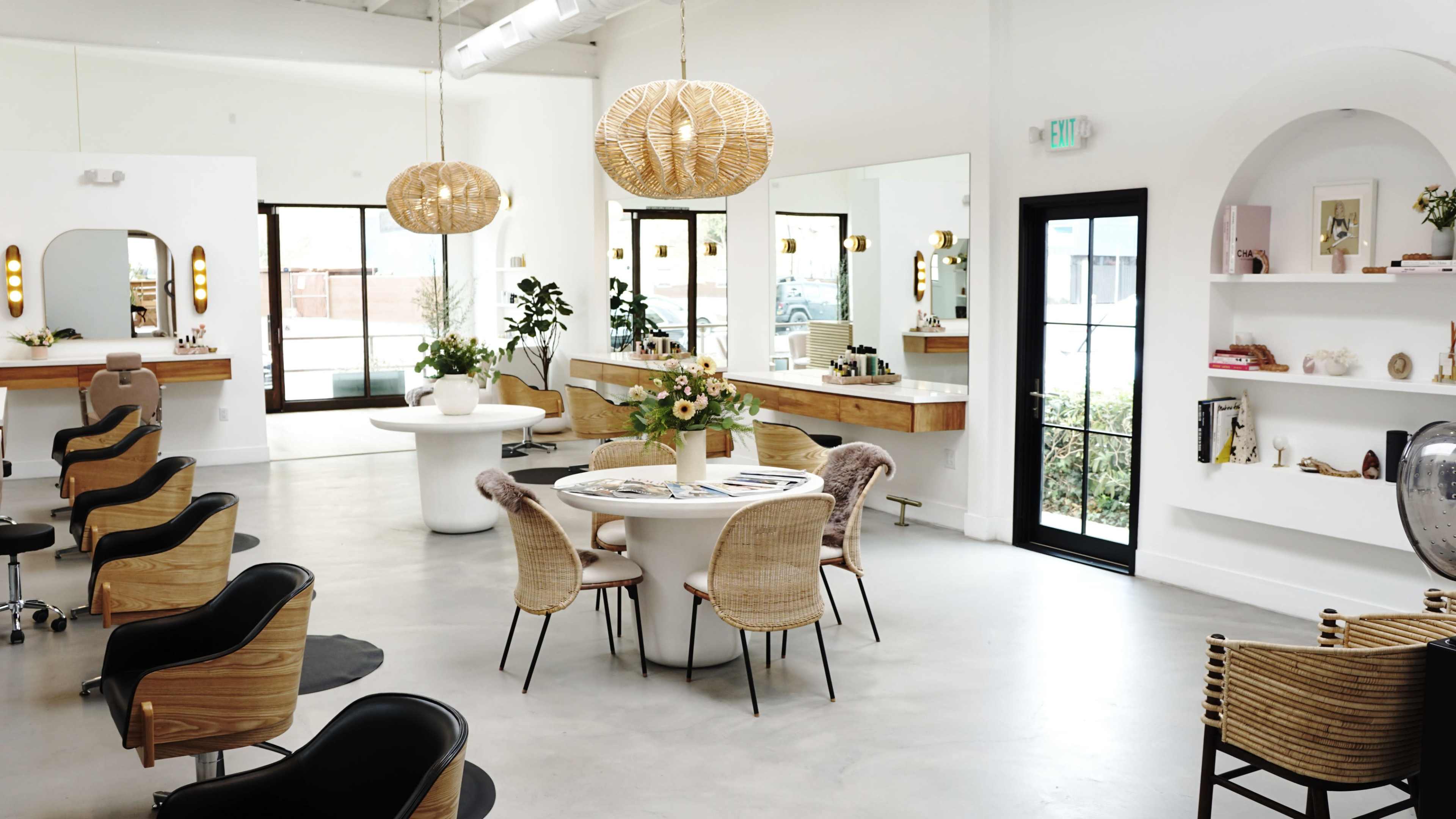 The image shows a modern, minimalist interior space featuring a round dining table surrounded by chairs, pendant lighting, and large windows with plants visible outside.