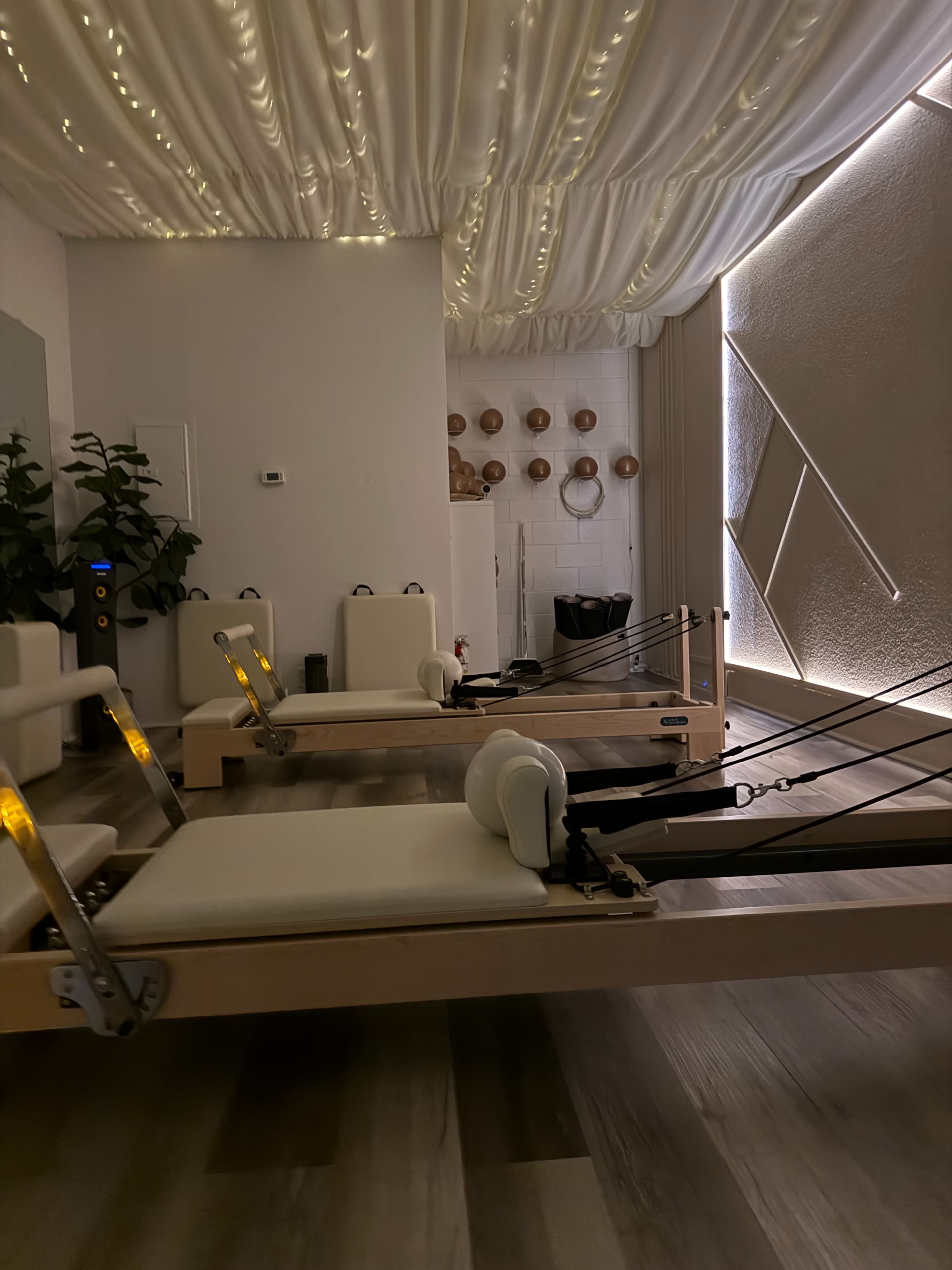 The image shows a tranquil room set up for Pilates, featuring several reformer machines arranged neatly under soft, ambient lighting and a draped ceiling.