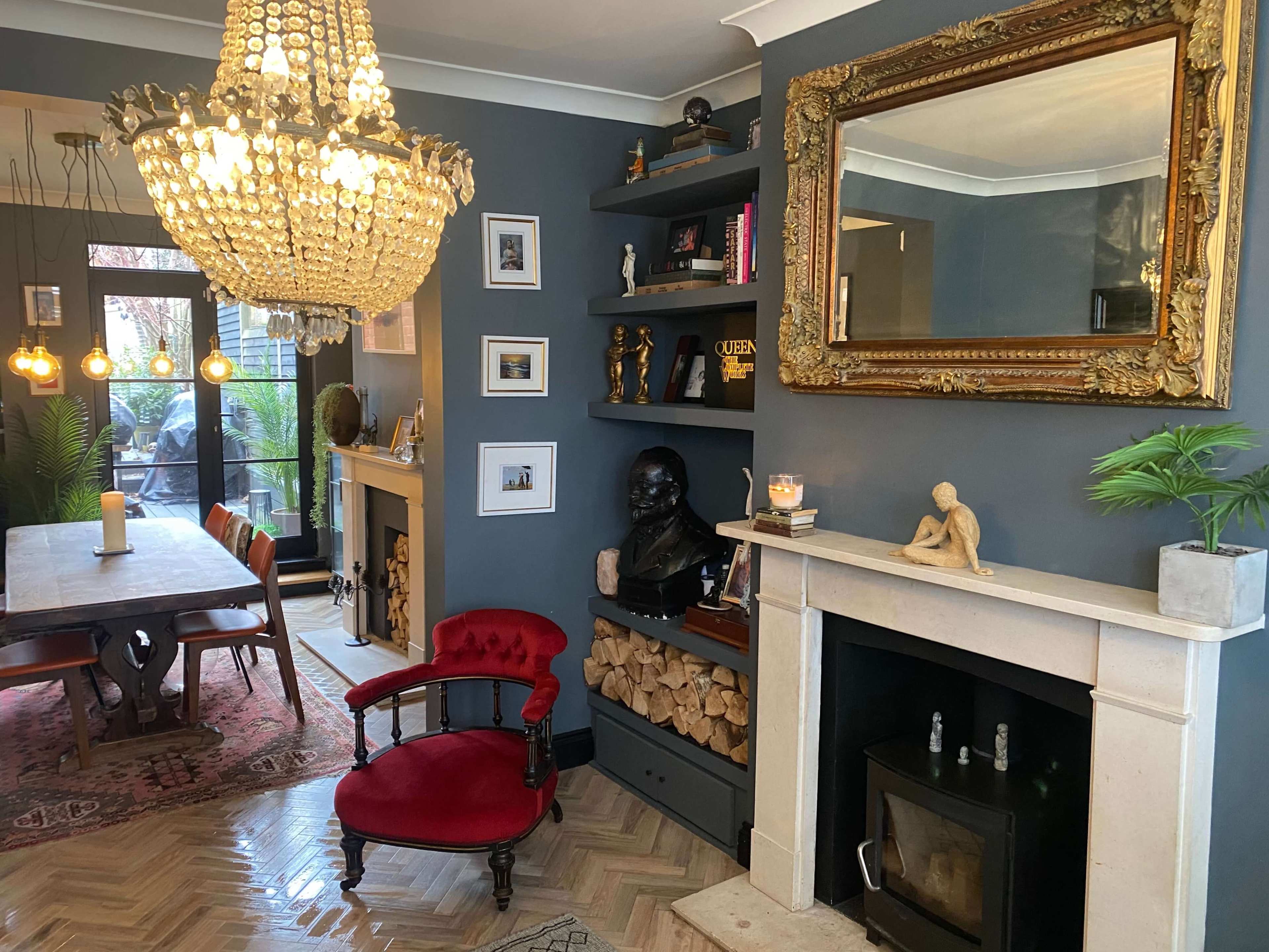 A stylish living room with a large chandelier, a wooden dining table, a fireplace, decorative shelves, and a vintage red armchair.