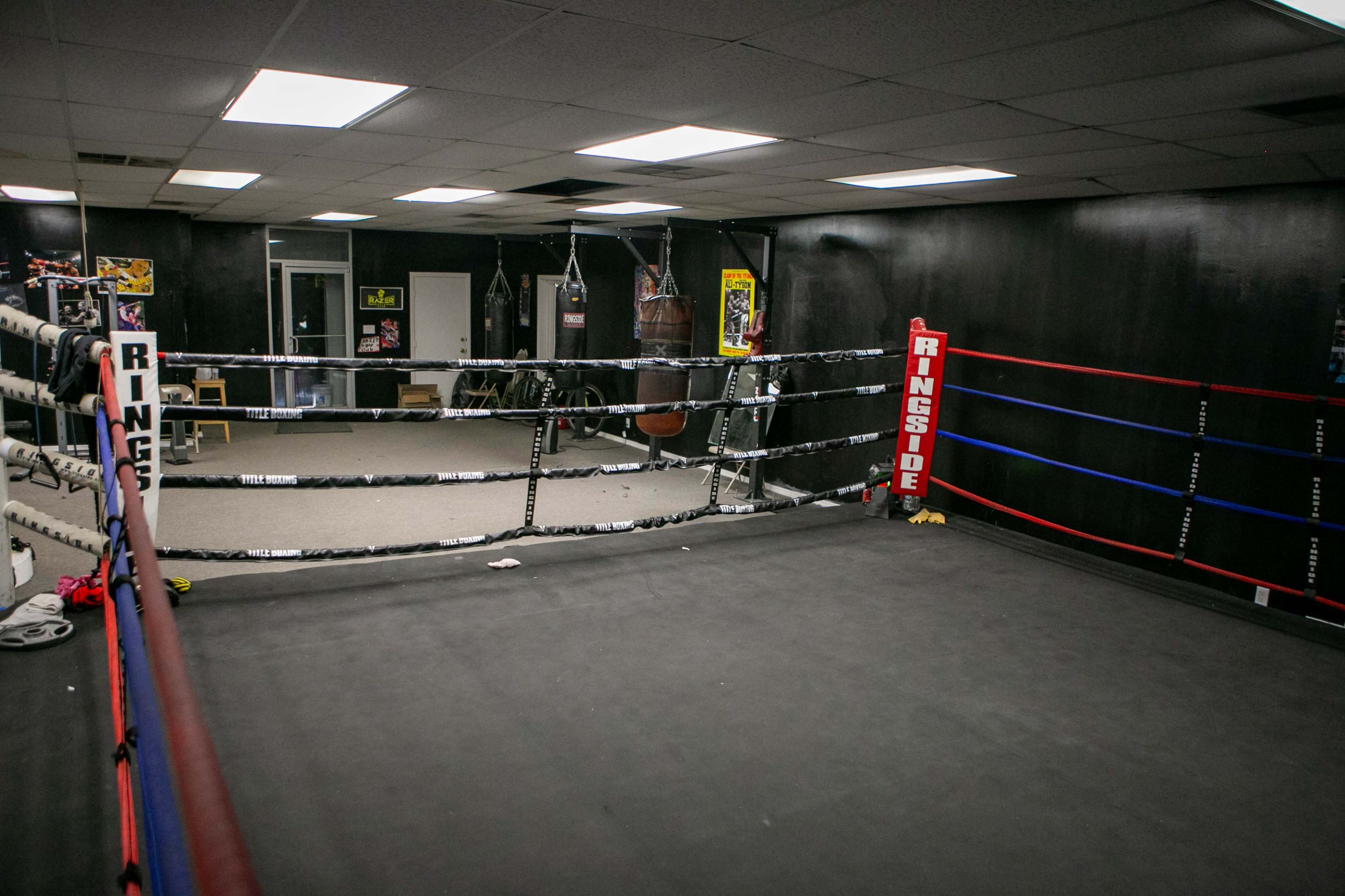The image shows an indoor boxing gym with a training ring surrounded by exercise equipment and heavy bags.