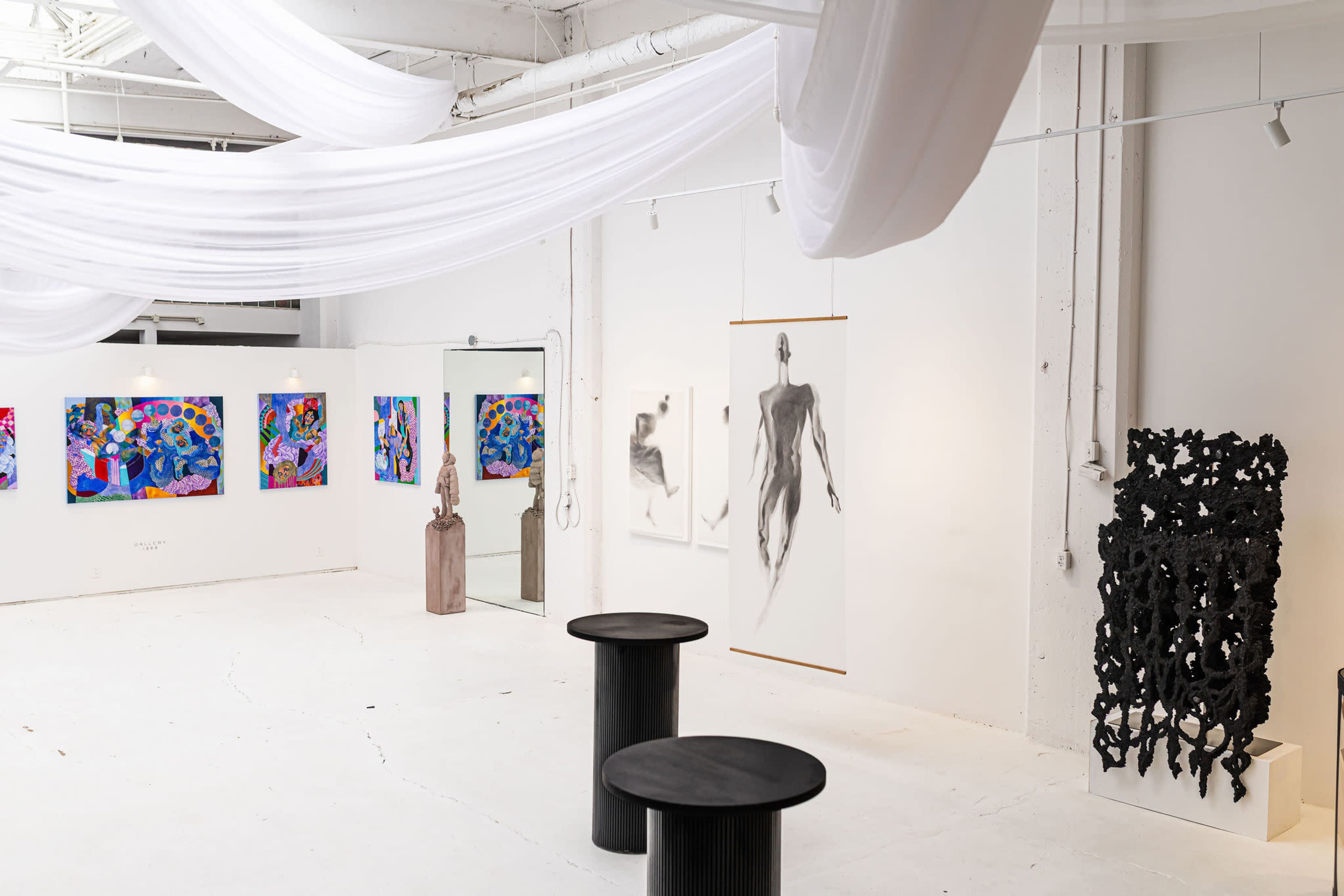 The image shows an art gallery featuring colorful paintings on the walls, a sculpture in the center, and two black round tables.