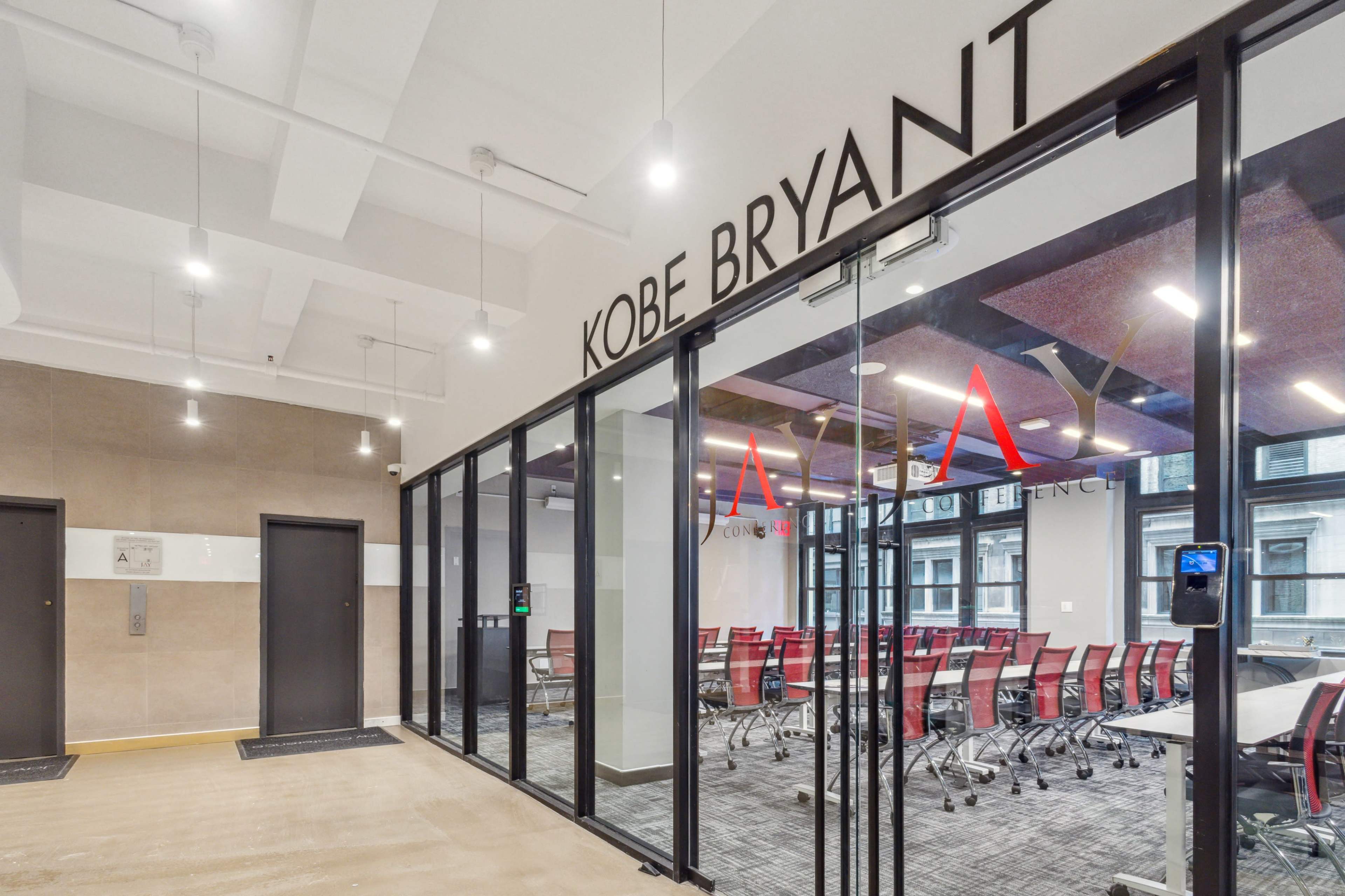 The image shows a modern conference room named "Kobe Bryant," featuring glass walls, red chairs, and a visible elevator area.