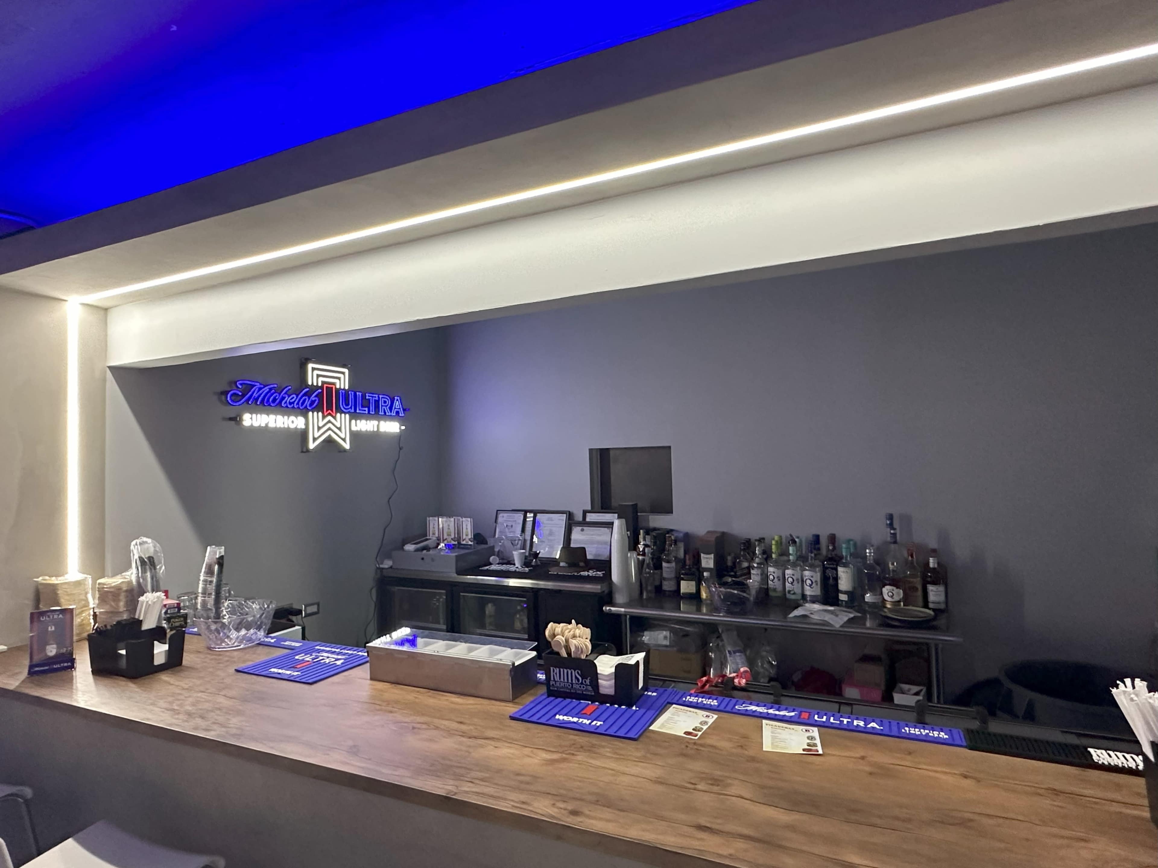 The image shows a modern bar counter with a selection of liquor bottles, glassware, and a neon Michelob Ultra sign in a well-lit space.