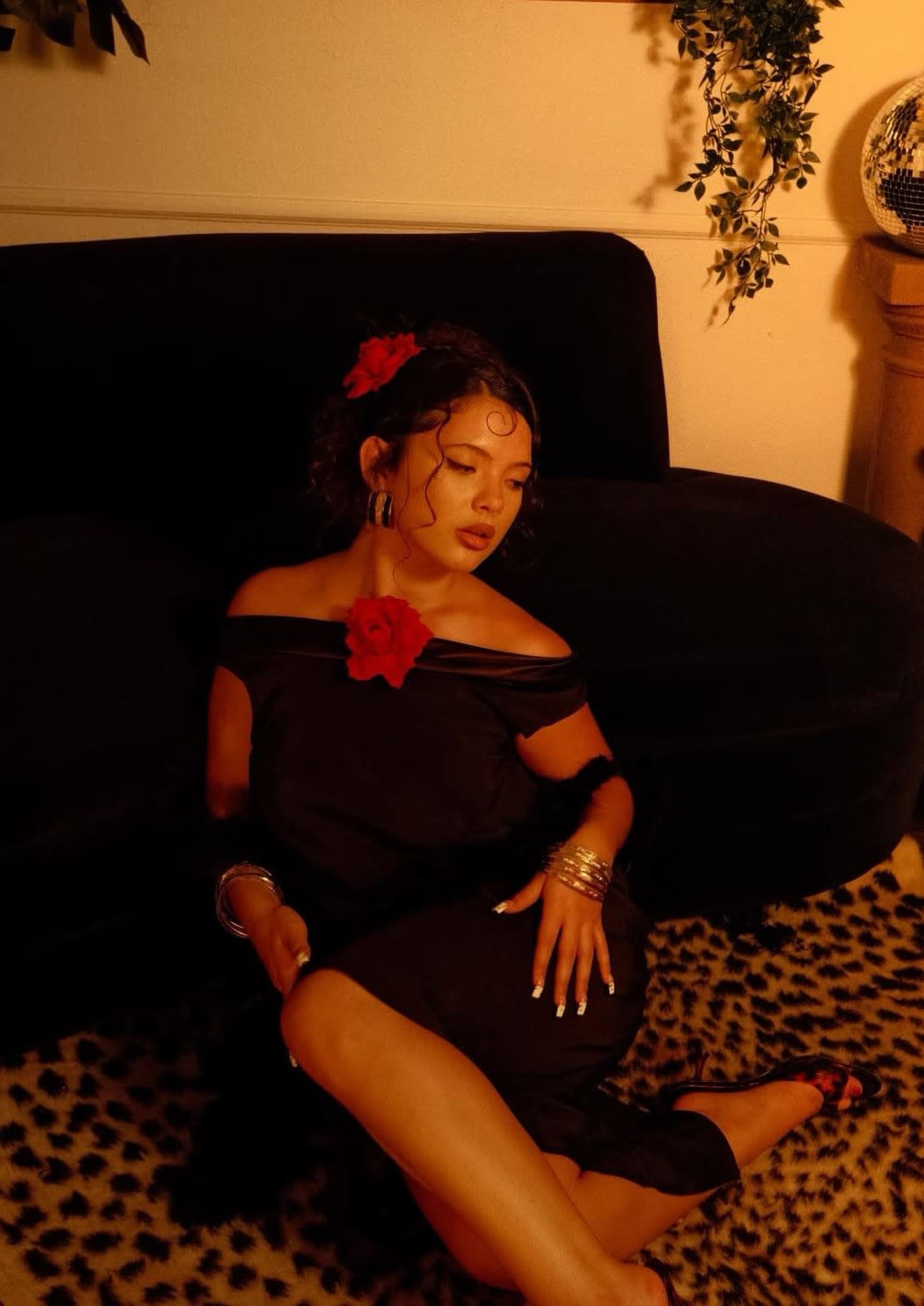 A person wearing a black dress and adorned with a red flower poses on a dark sofa against a backdrop of soft lighting and animal print.