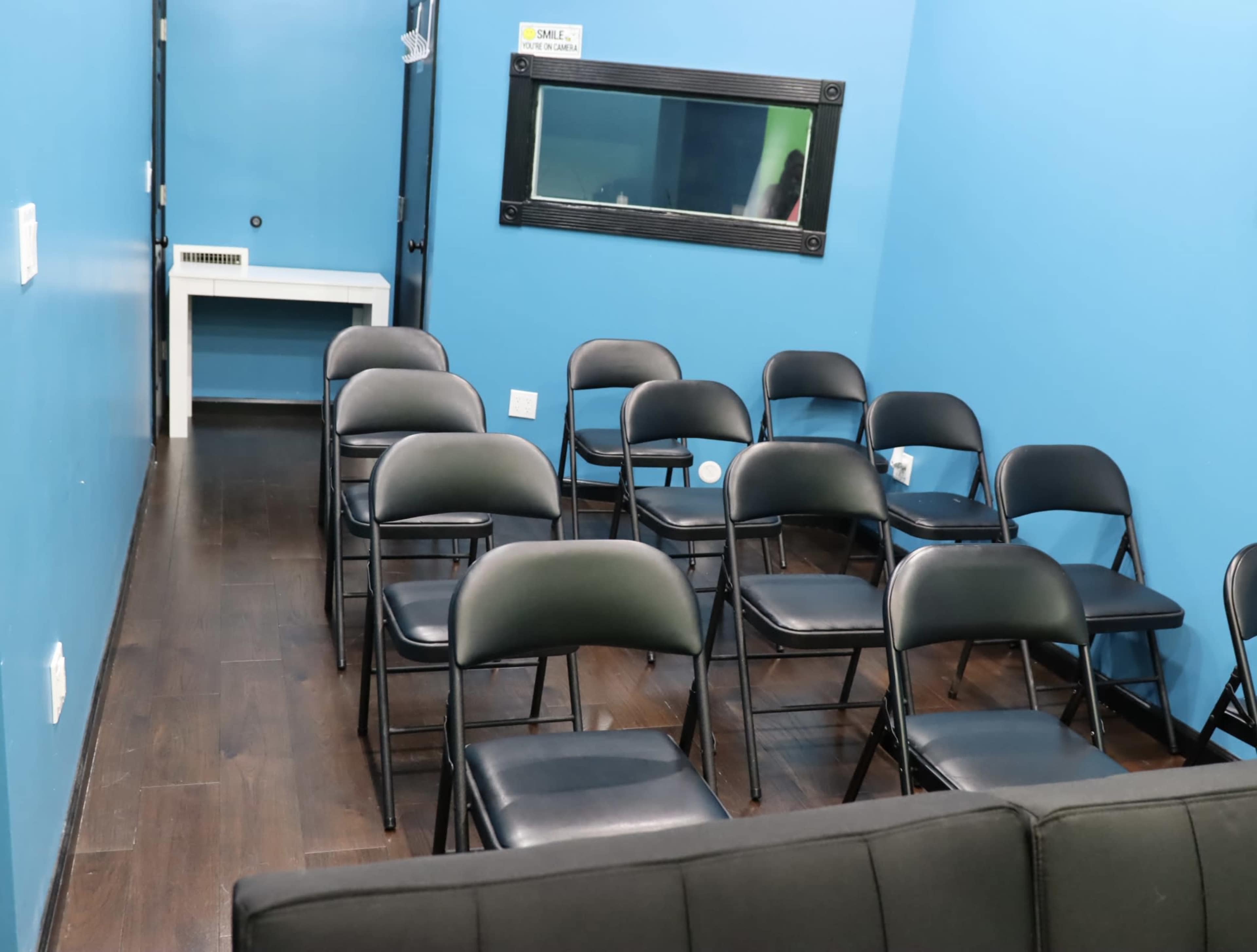 A small, blue-walled room with several black folding chairs arranged in rows facing a wall mirror.