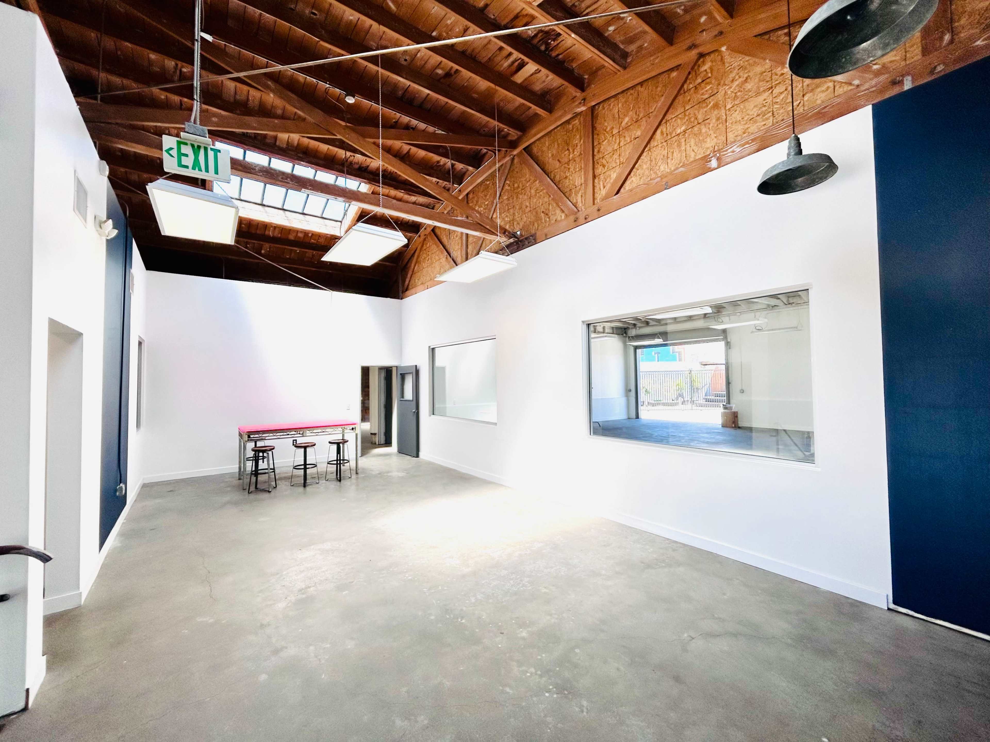 The image shows a spacious, well-lit room with a high ceiling, featuring exposed beams, concrete flooring, and a table with stools.