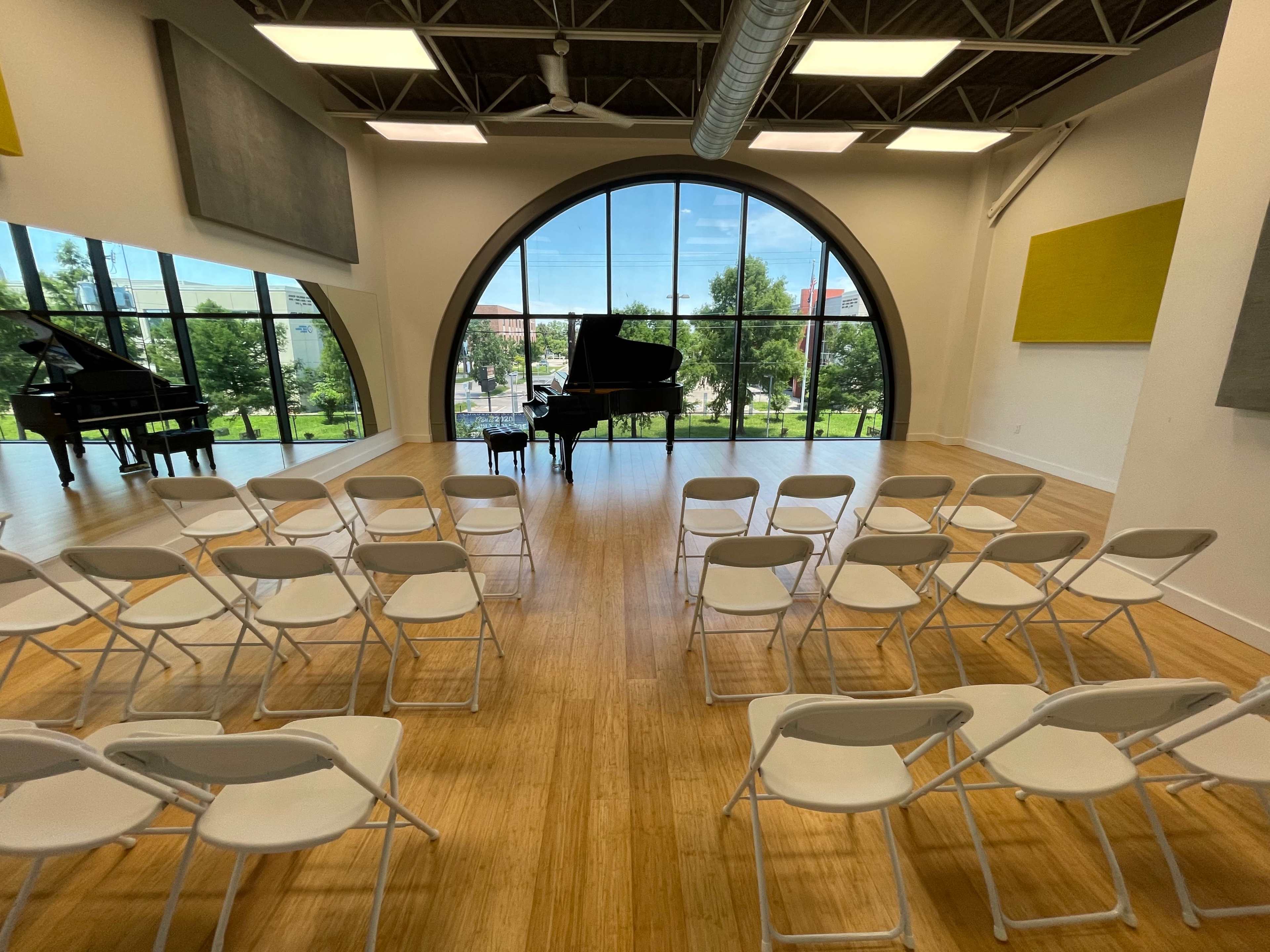 The image shows a spacious music room with two grand pianos and white folding chairs arranged in front of them, set against large windows that overlook a green landscape.