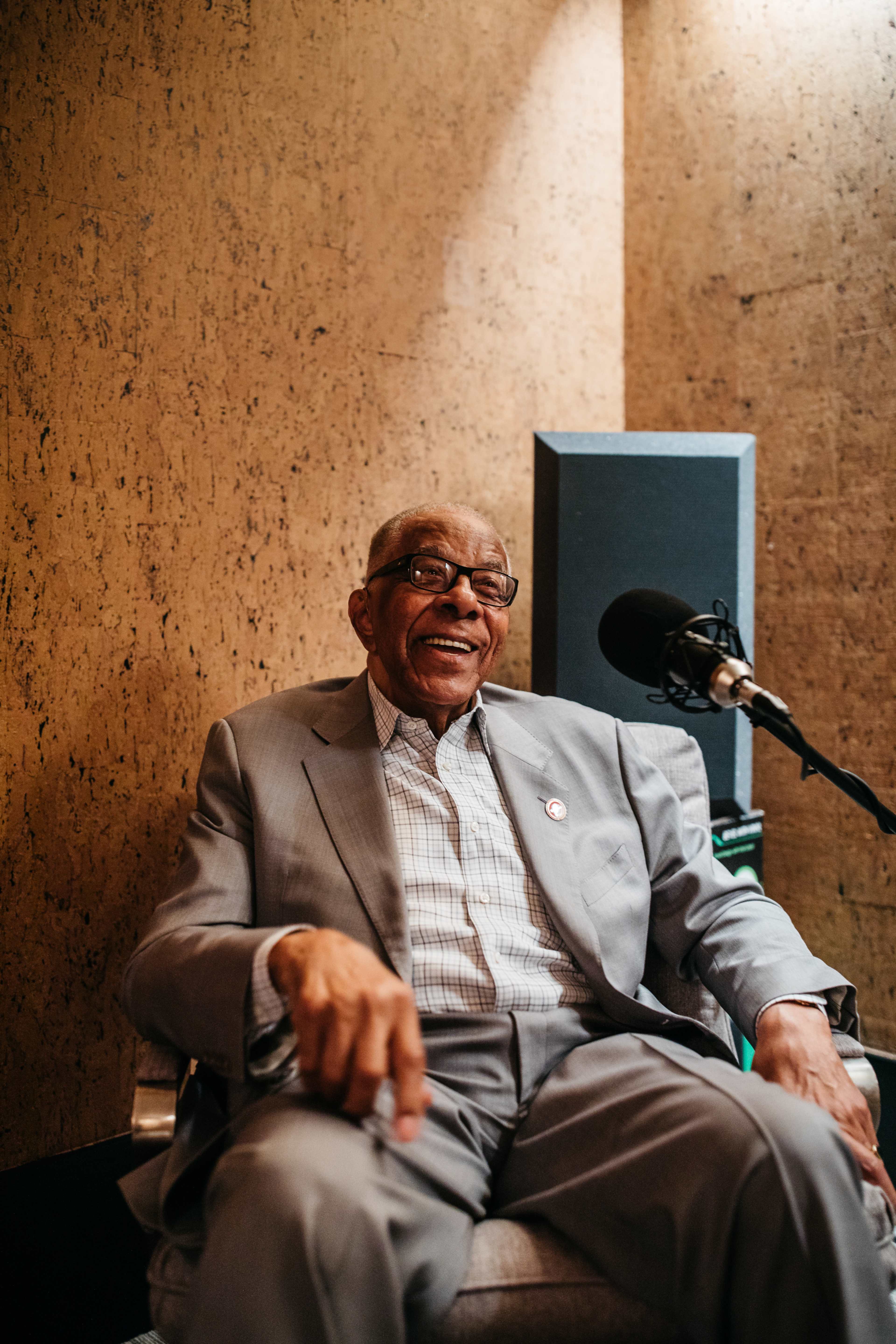 An older man in a gray suit sits comfortably in a chair with a microphone nearby, smiling against a textured wall.