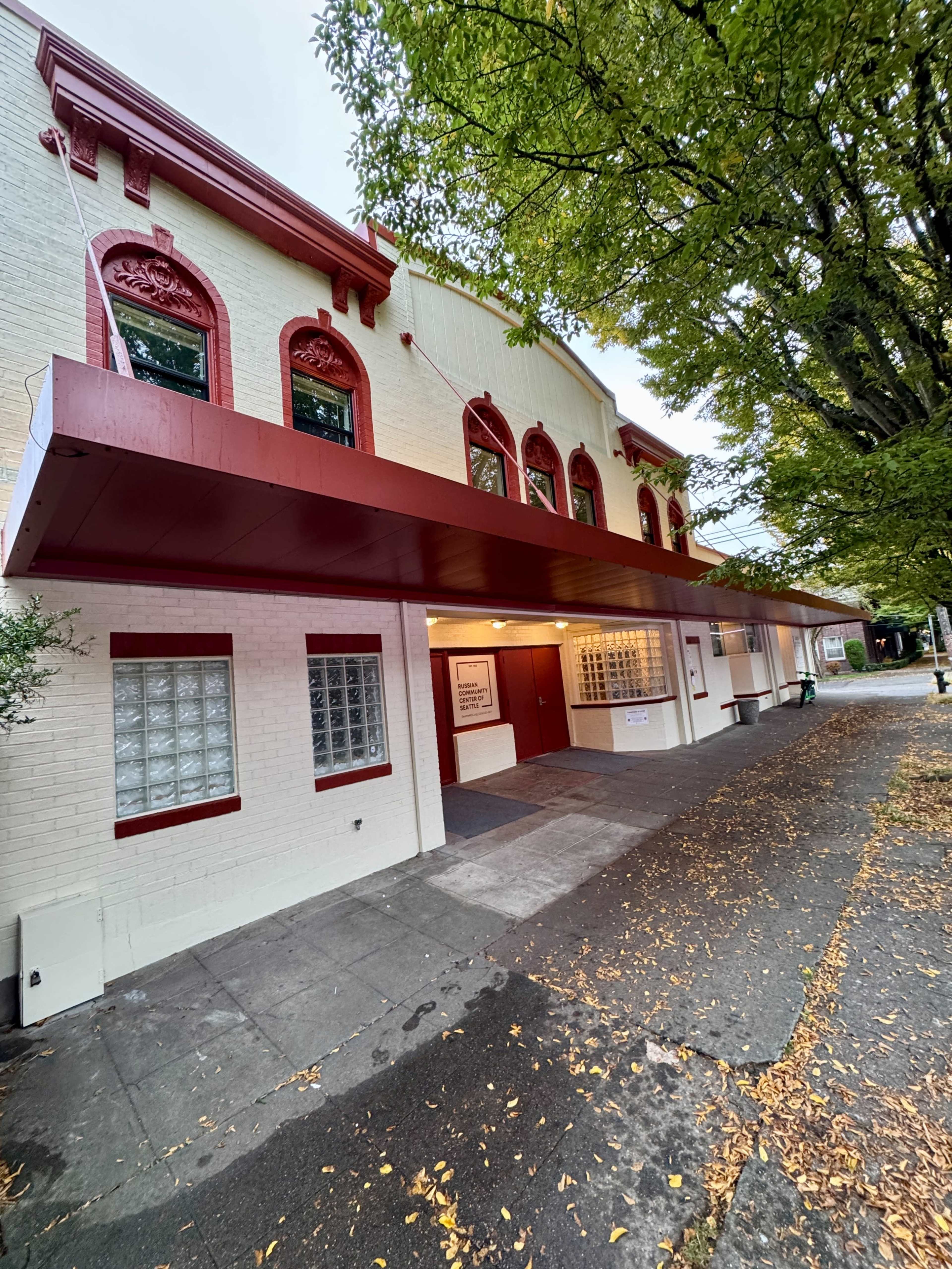 The image shows a building with a red canopy and decorative archways, surrounded by trees and fallen leaves on the sidewalk.