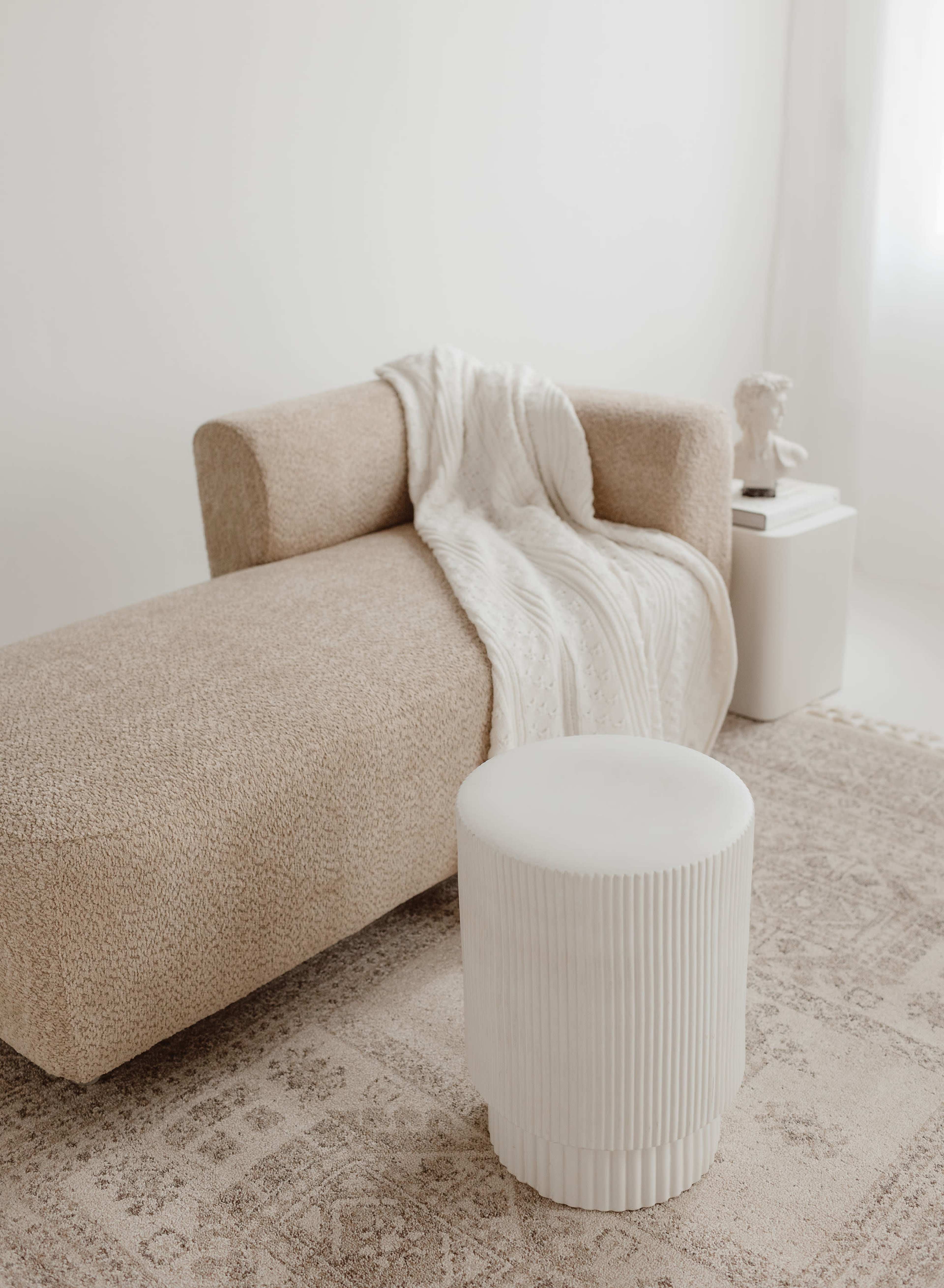 A textured beige sofa with a blanket draped over it is positioned next to a white, cylindrical side table on a patterned rug.