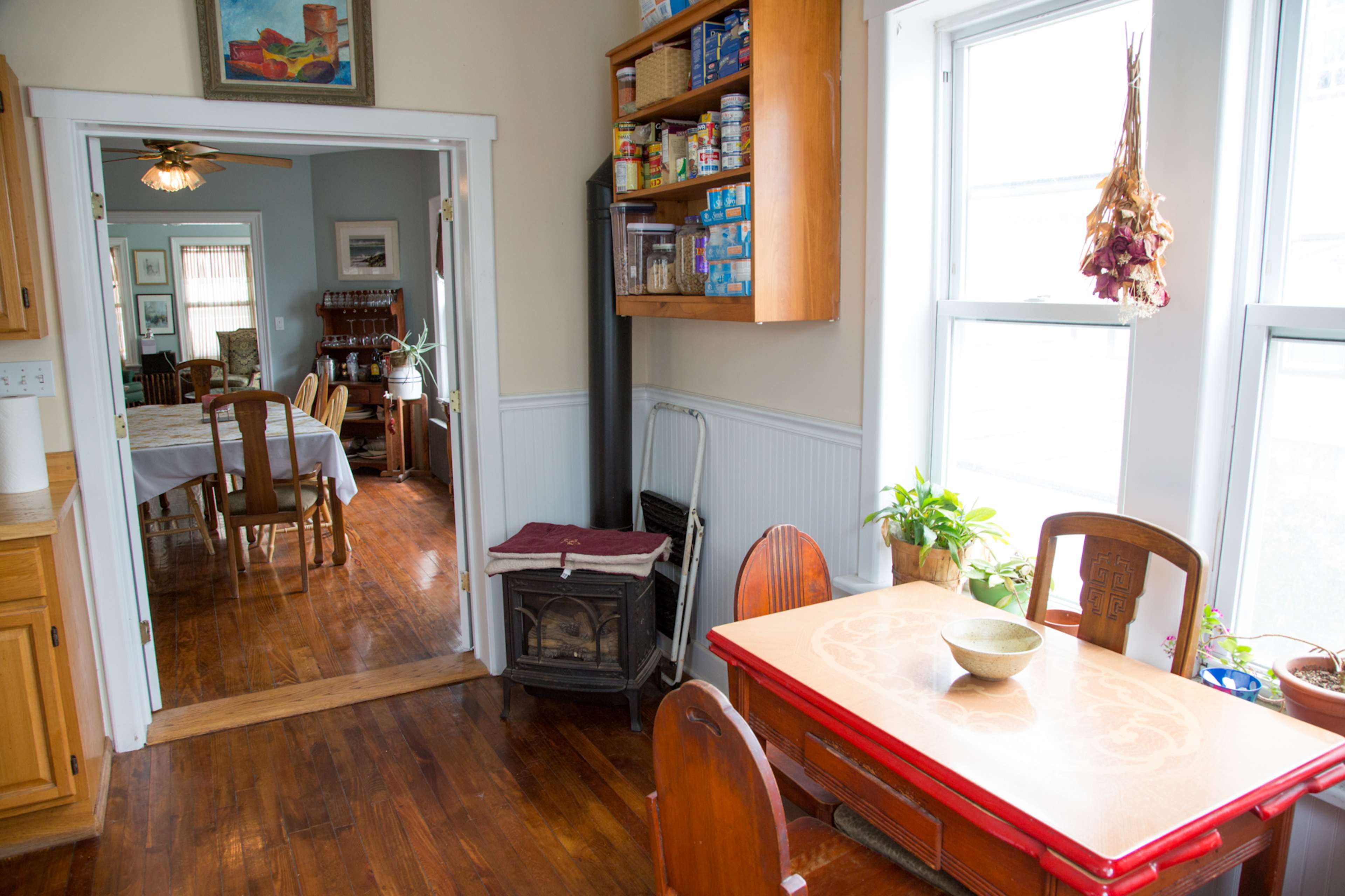 The image shows a bright kitchen with a red table and wooden chairs, opening into a dining area with a table set for a meal.