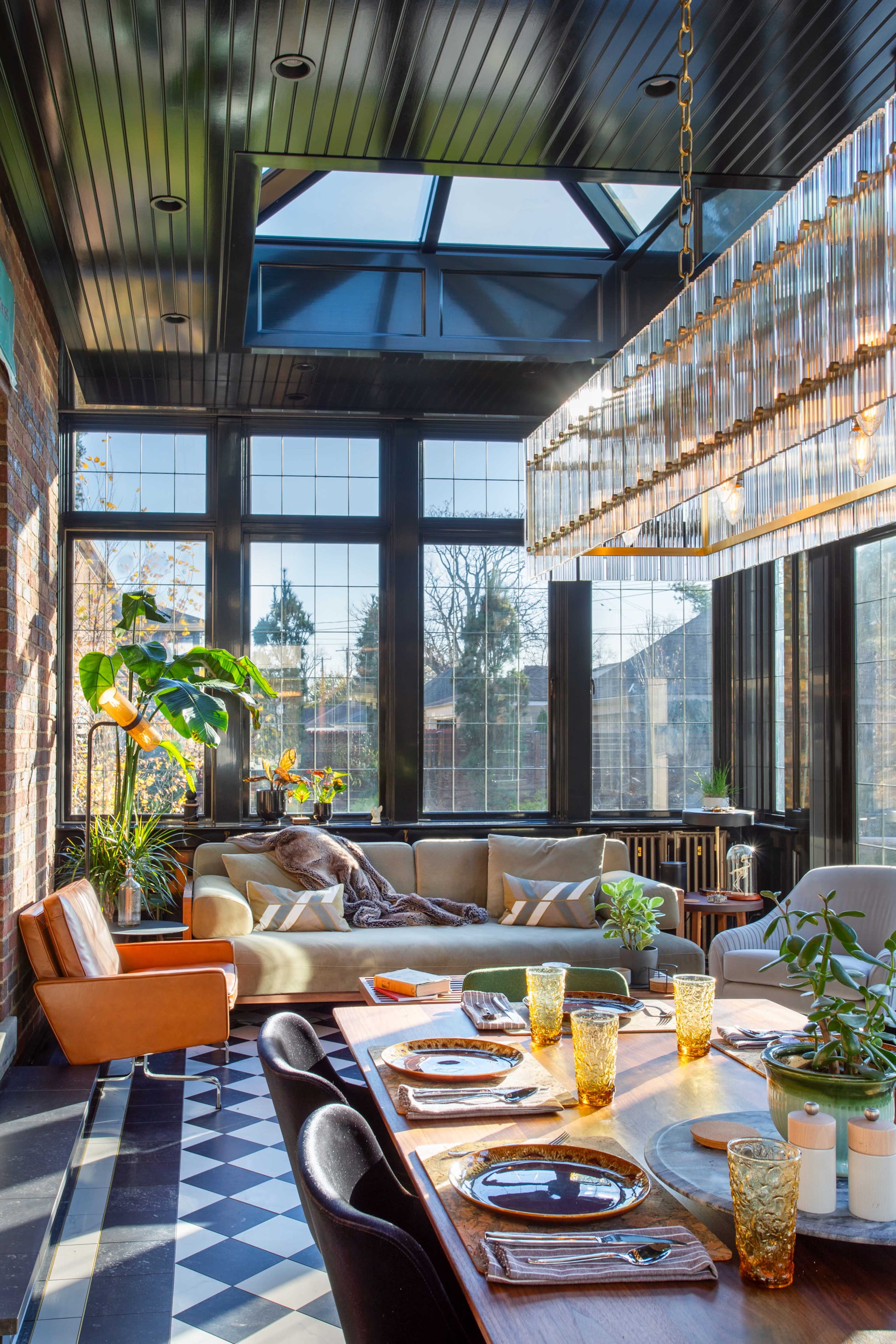 The image shows a bright sunroom with large windows, a dining table set with yellow glasses, and a mix of seating including a couch and armchair.