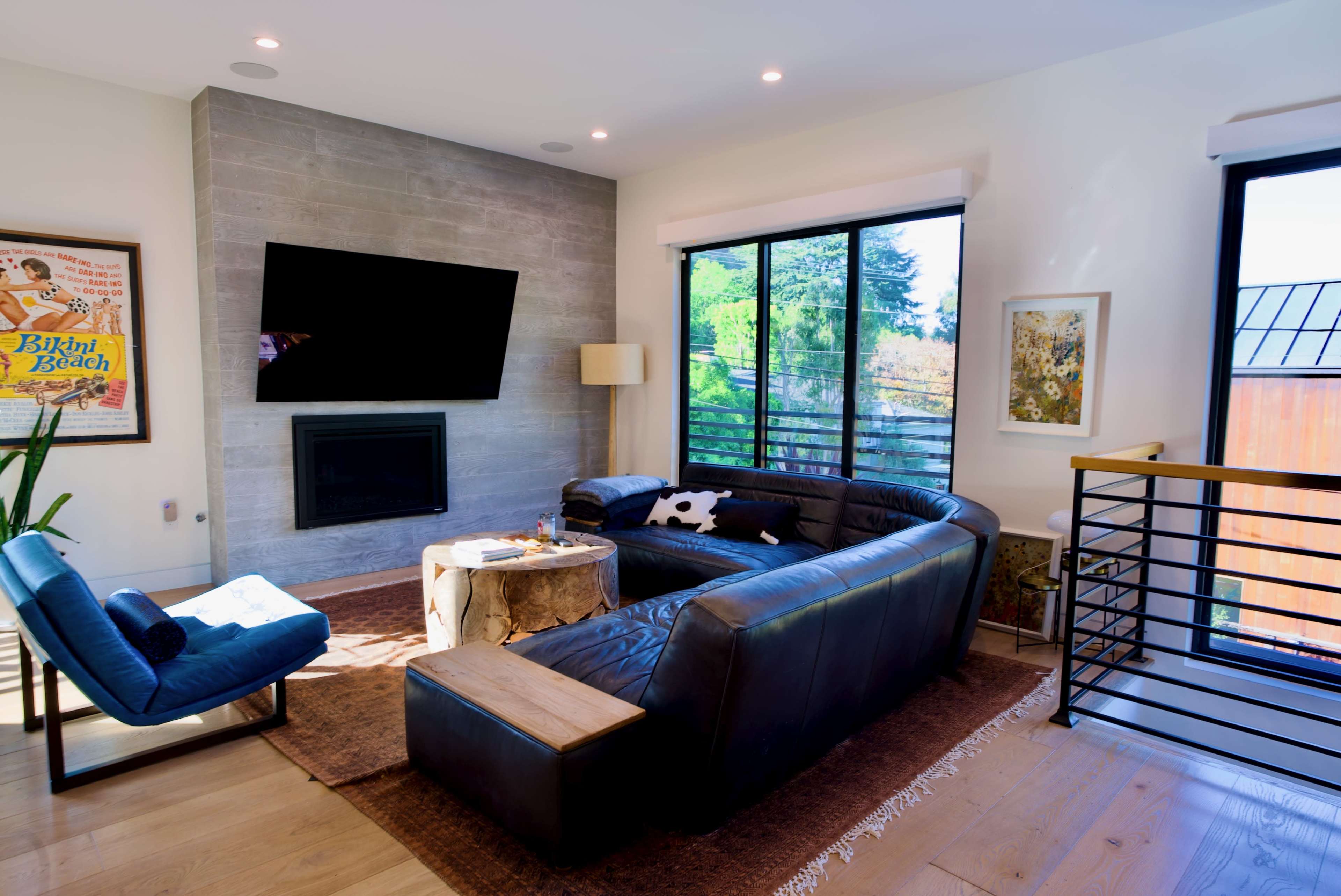 A modern living room features a large flat-screen TV mounted on a concrete wall, a dark sectional sofa, and a cozy armchair beside a window with natural light.