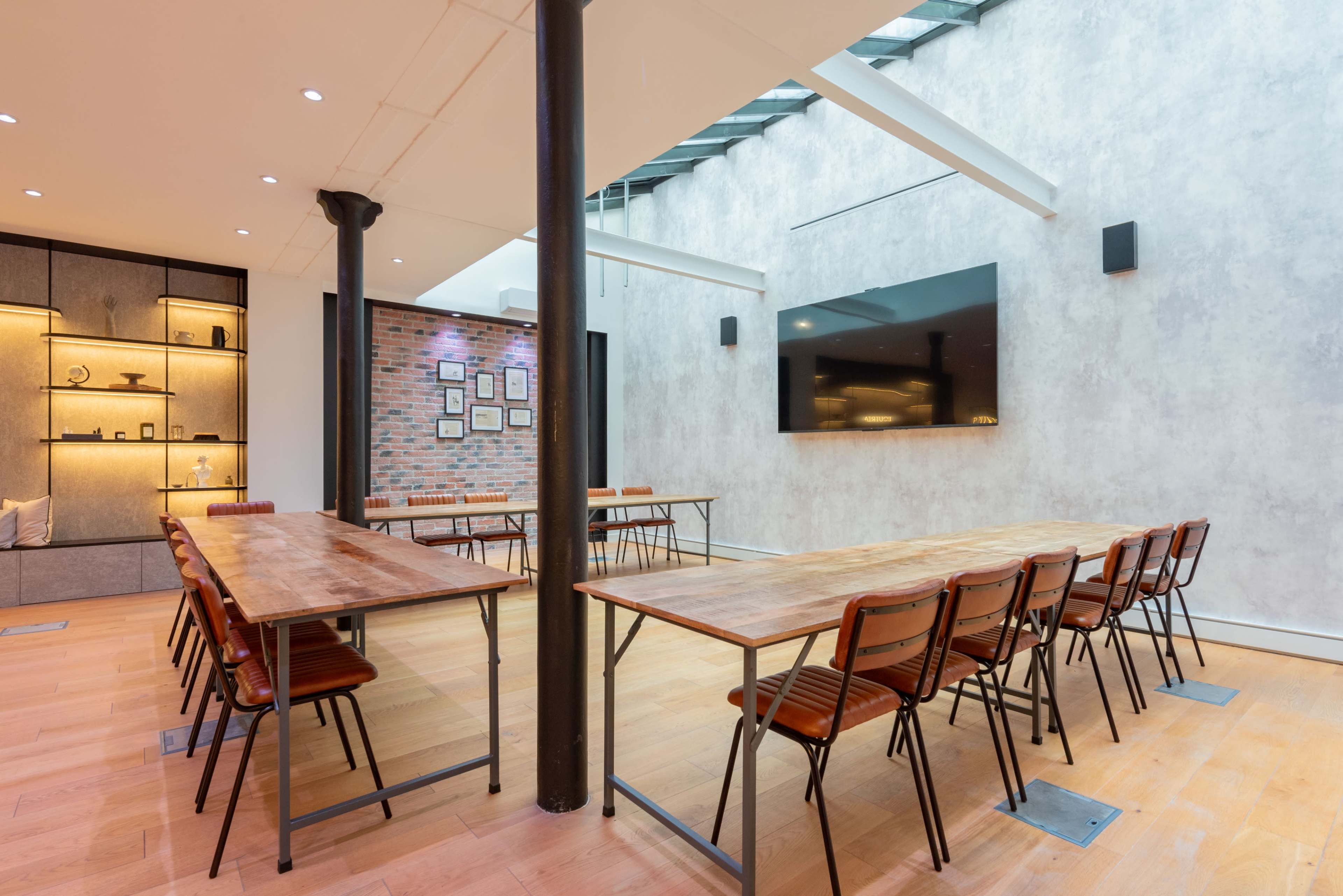 The image shows a modern conference room featuring multiple wooden tables arranged in a U-shape, with chairs and a large screen mounted on a gray wall.