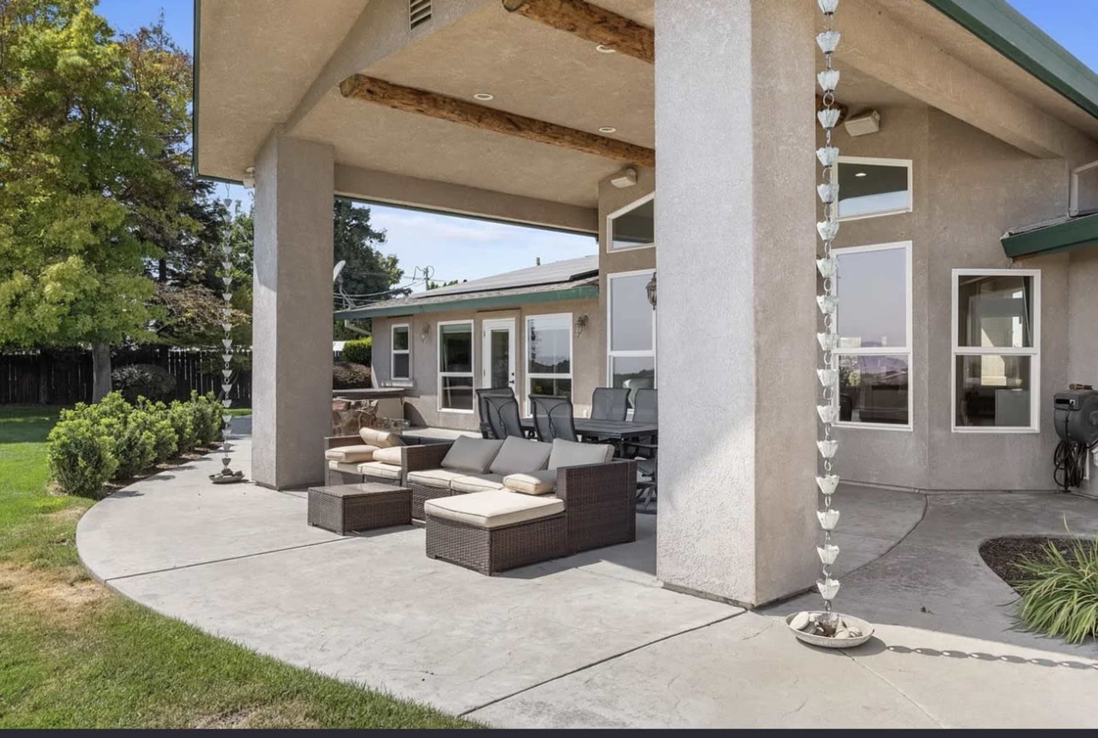 The image shows a patio area with outdoor seating and a concrete pathway, adjacent to a house surrounded by a lawn and trees.