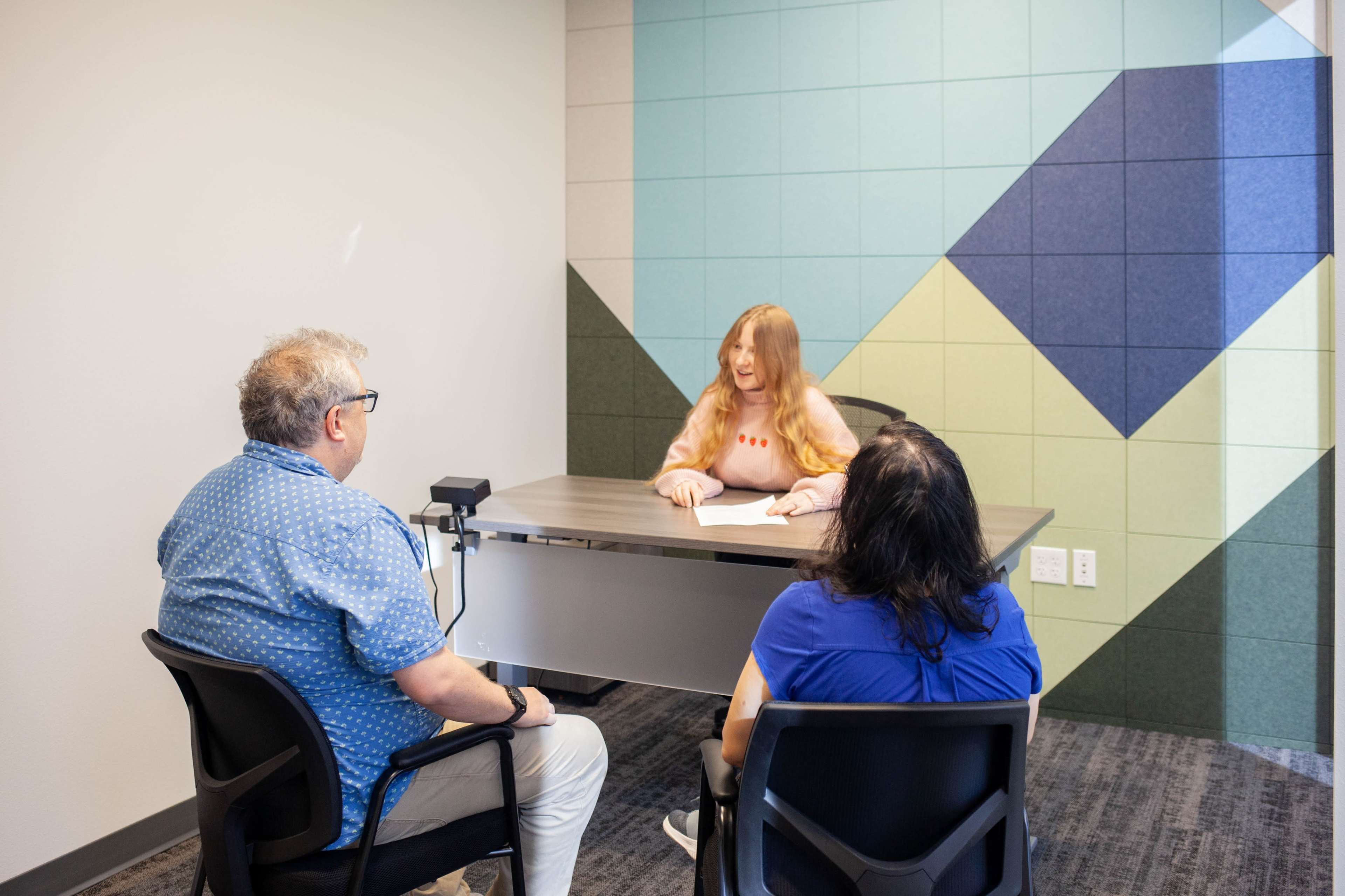 A woman with long hair sits at a table facing two seated individuals in a brightly colored room with geometric wall designs.