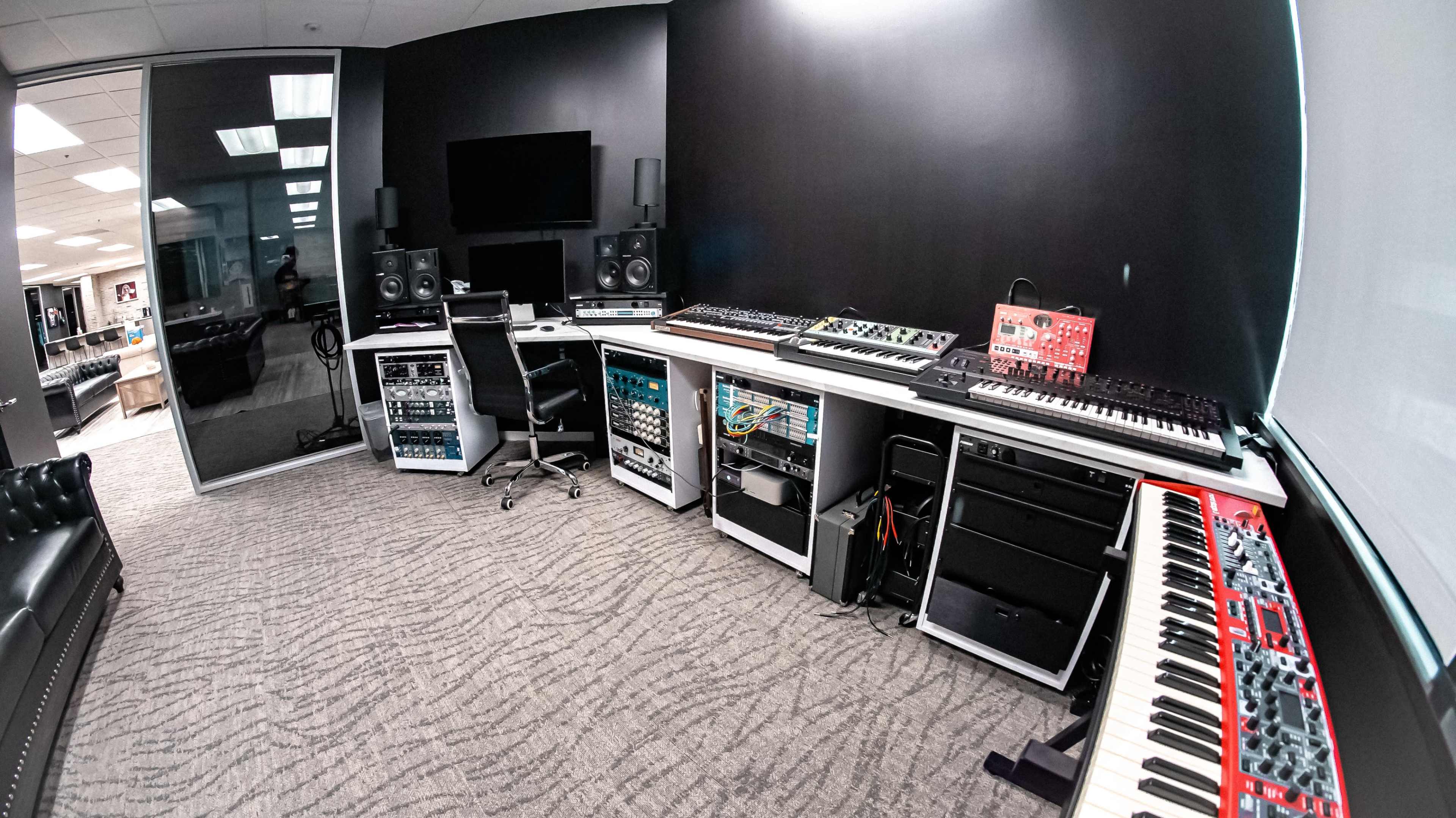 A modern music production studio featuring multiple synthesizers, audio equipment, and a computer workstation arranged around a central area with a couch.