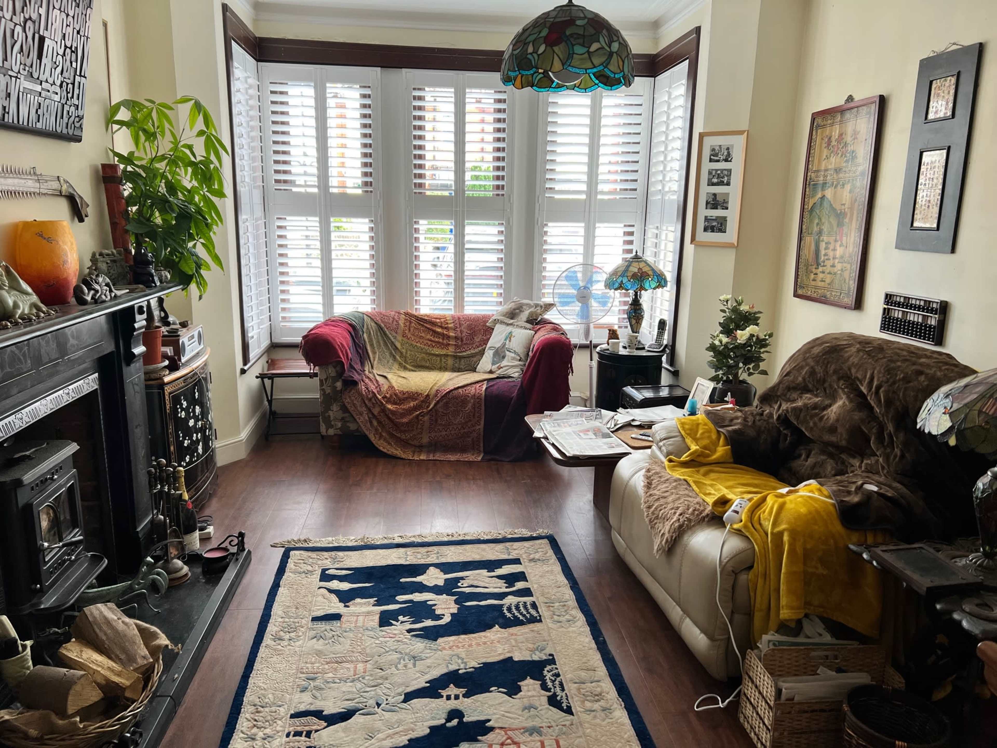 A cozy living room features a red couch, a white sofa covered in yellow and brown blankets, a patterned rug, and plants beside a decorative fireplace.