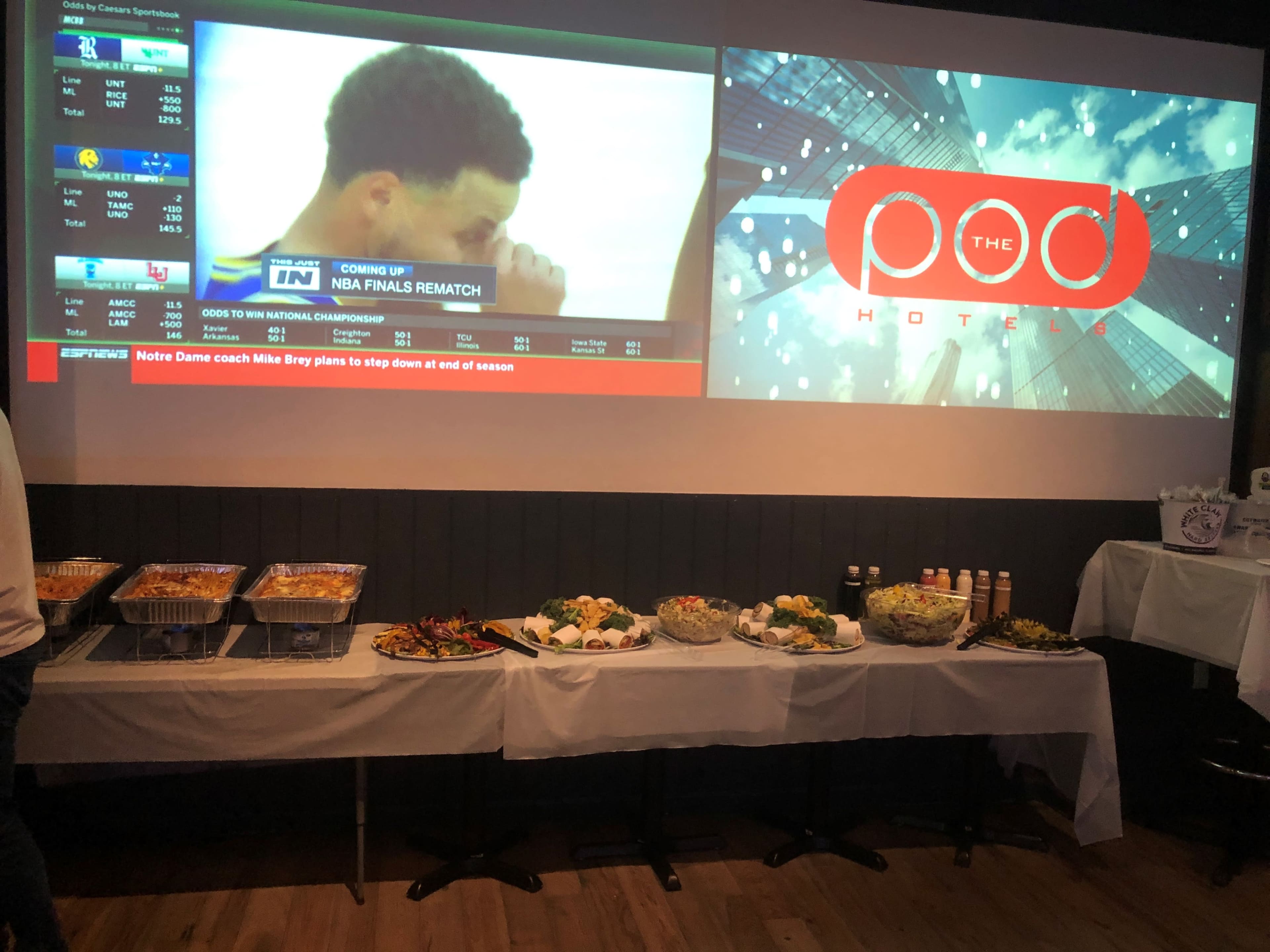 A buffet table with various dishes is set up in front of a large screen displaying sports highlights and promotional content for a hotel.